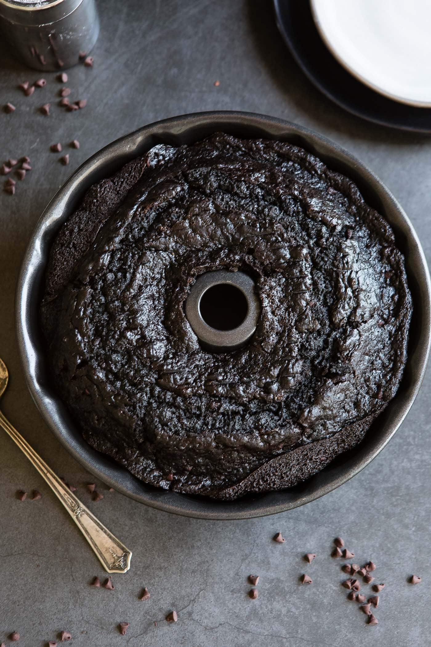 A baked zucchini chocolate bundt cake in a bundt pan. A cake server and mini chocolate chips are on the table next to the cake.