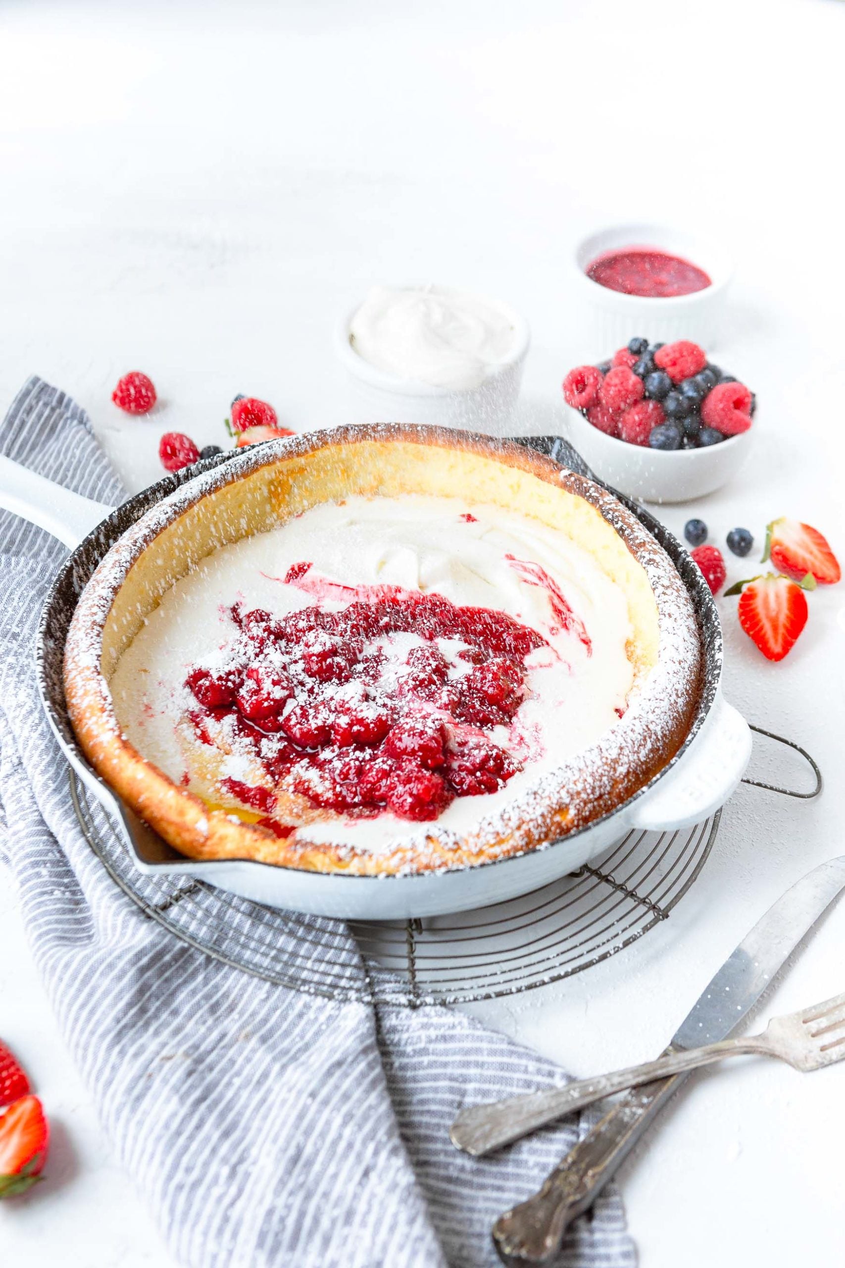 A photo of a baked dutch baby pancake in a cast iron skillet topped with whipped cream, fresh berries and powdered sugar.