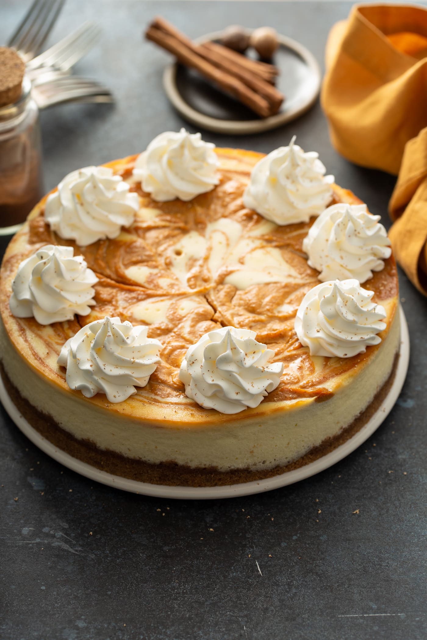 A whole swirled pumpkin cheesecake topped with dollops of whipped cream. There is a napkin, cinnamon sticks and whole nutmeg in the background.