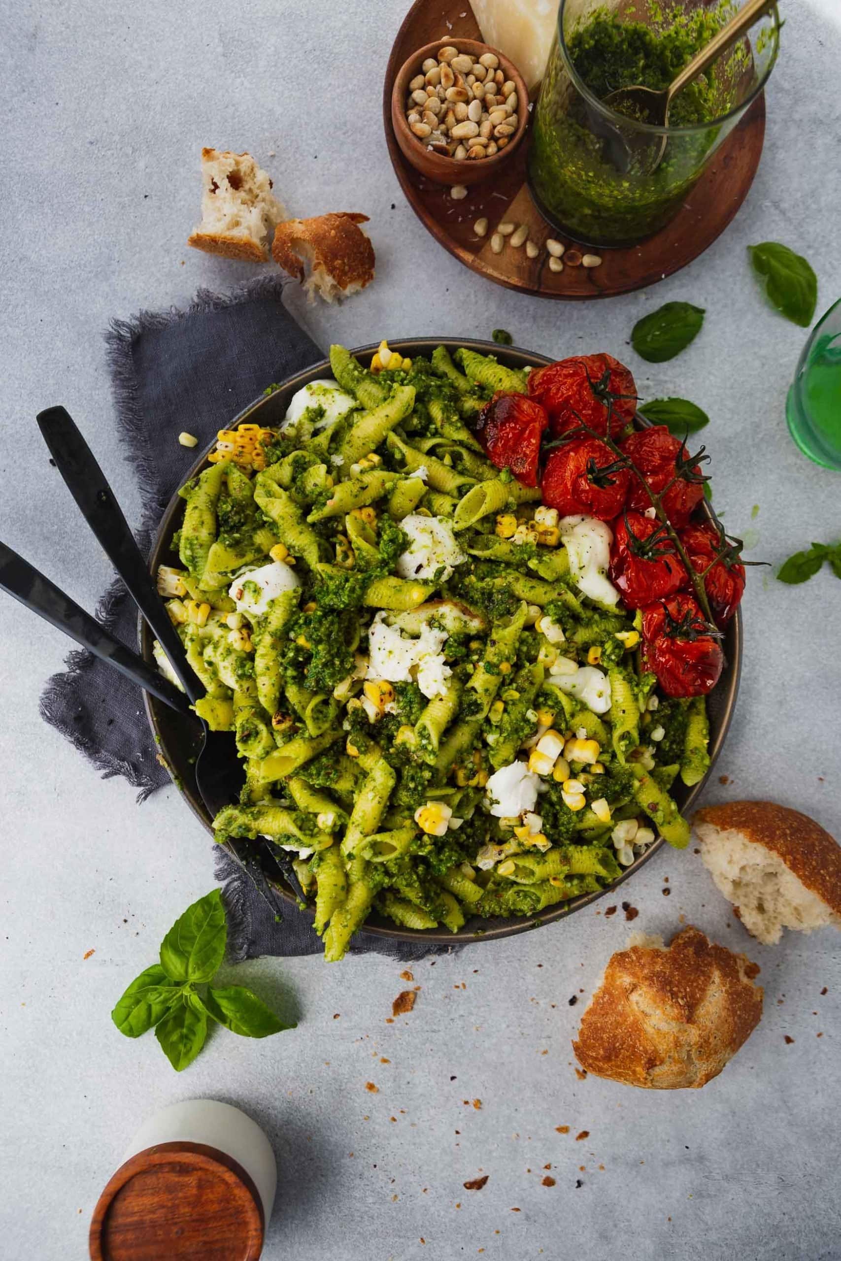 A photo of a large bowl full of pesto pasta topped with roasted tomatoes, grilled corn and mozzarella balls with serving spoons stuck in the bowl.