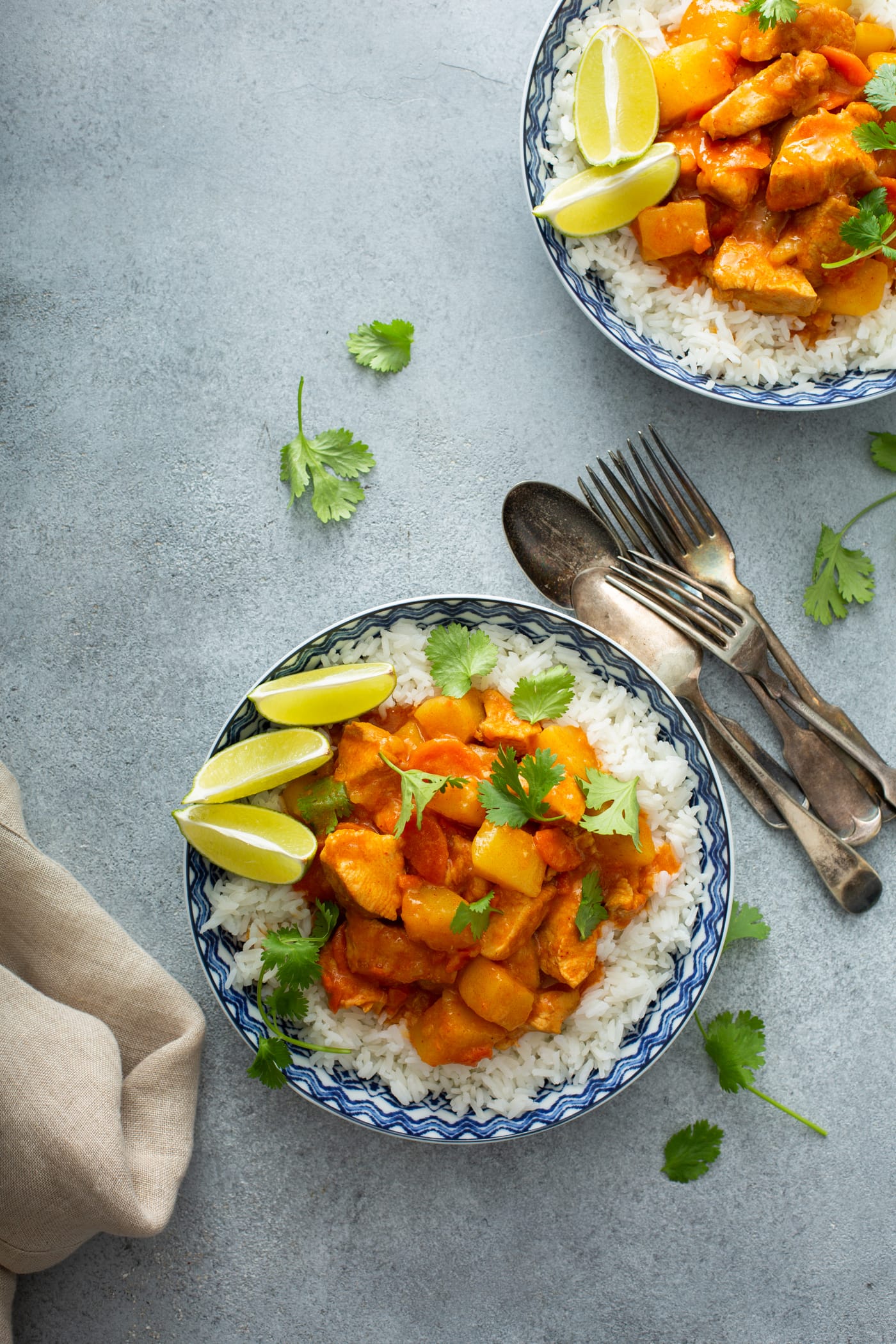 A bowl of rice topped with chicken curry. There are lime wedges on the edge of the bowl and forks and spoons next to the bowl. Another bowl of curry is in the background.