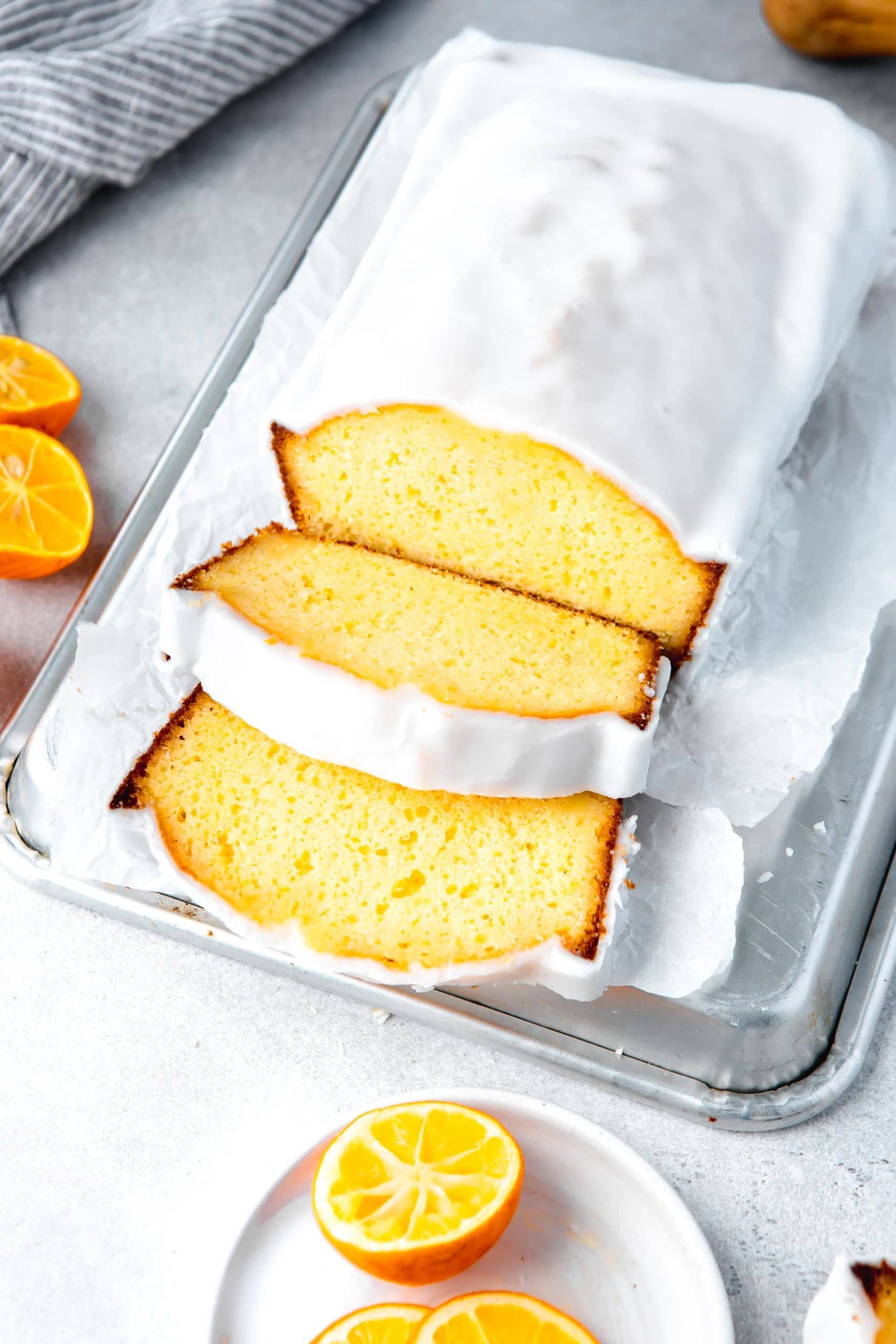 a photo of an iced lemon loaf cake sitting on a small silver baking sheet on top of a piece of parchment paper. Two slices have been cut off the cake and are laying next to the loaf.