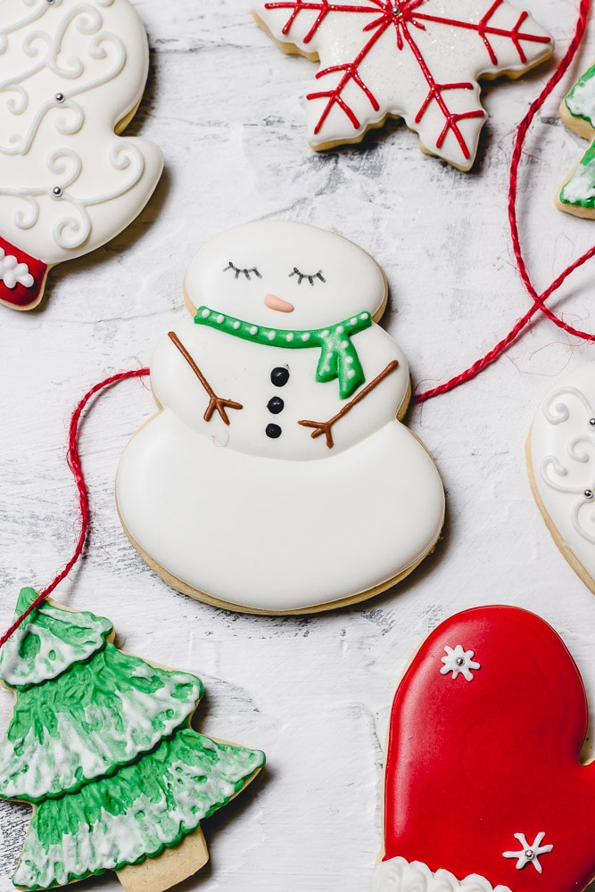 Christmas sugar cookies decorated with royal icing. There is a snowman with a green scarf, a green tree with white snow, a red mitten and a white mitten.