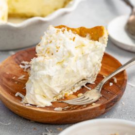 a photo of a slice of coconut cream pie topped with toasted shredded coconut flakes on a small wooden plate with a fork sitting next to the slice of pie.