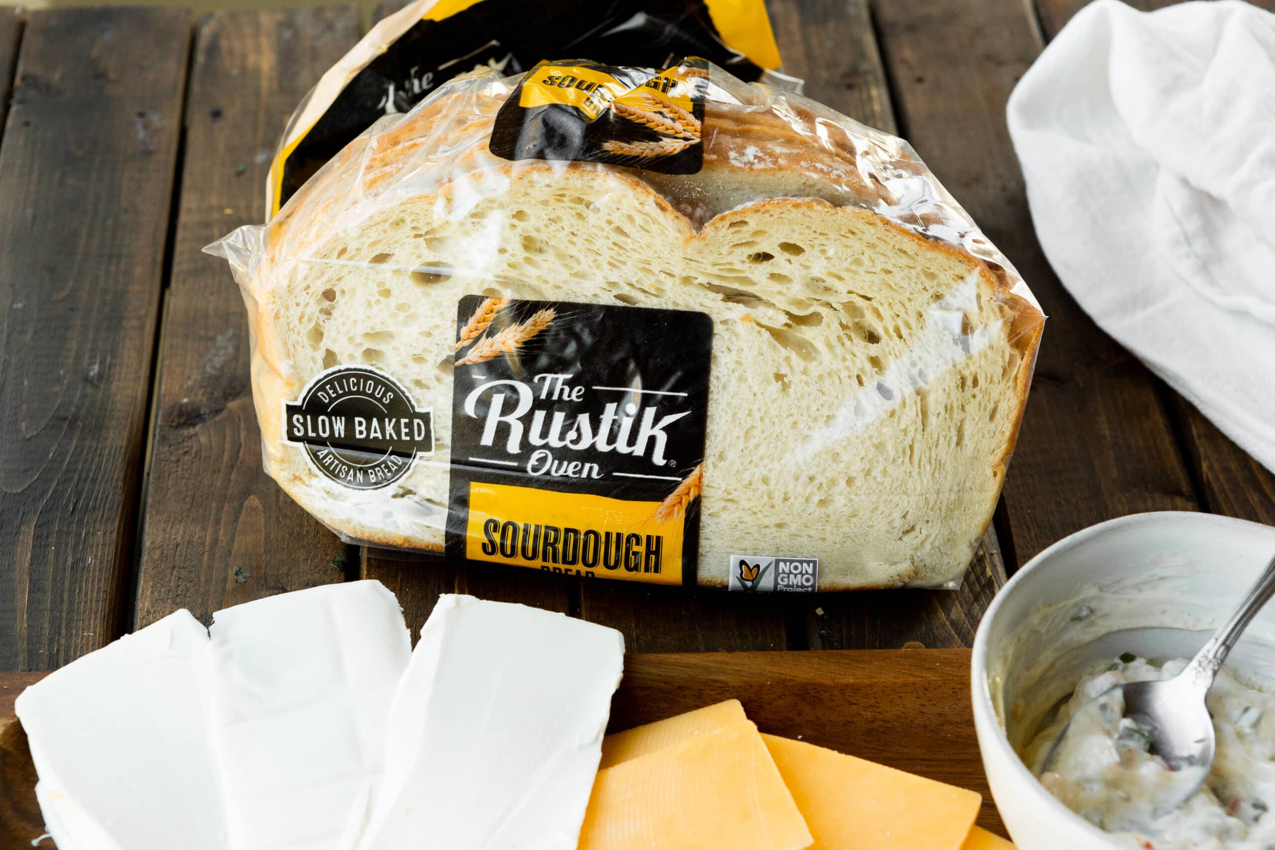 a photo of a bag of The Rustik Oven sourdough bread with slices of two different types of cheese laying next to it.