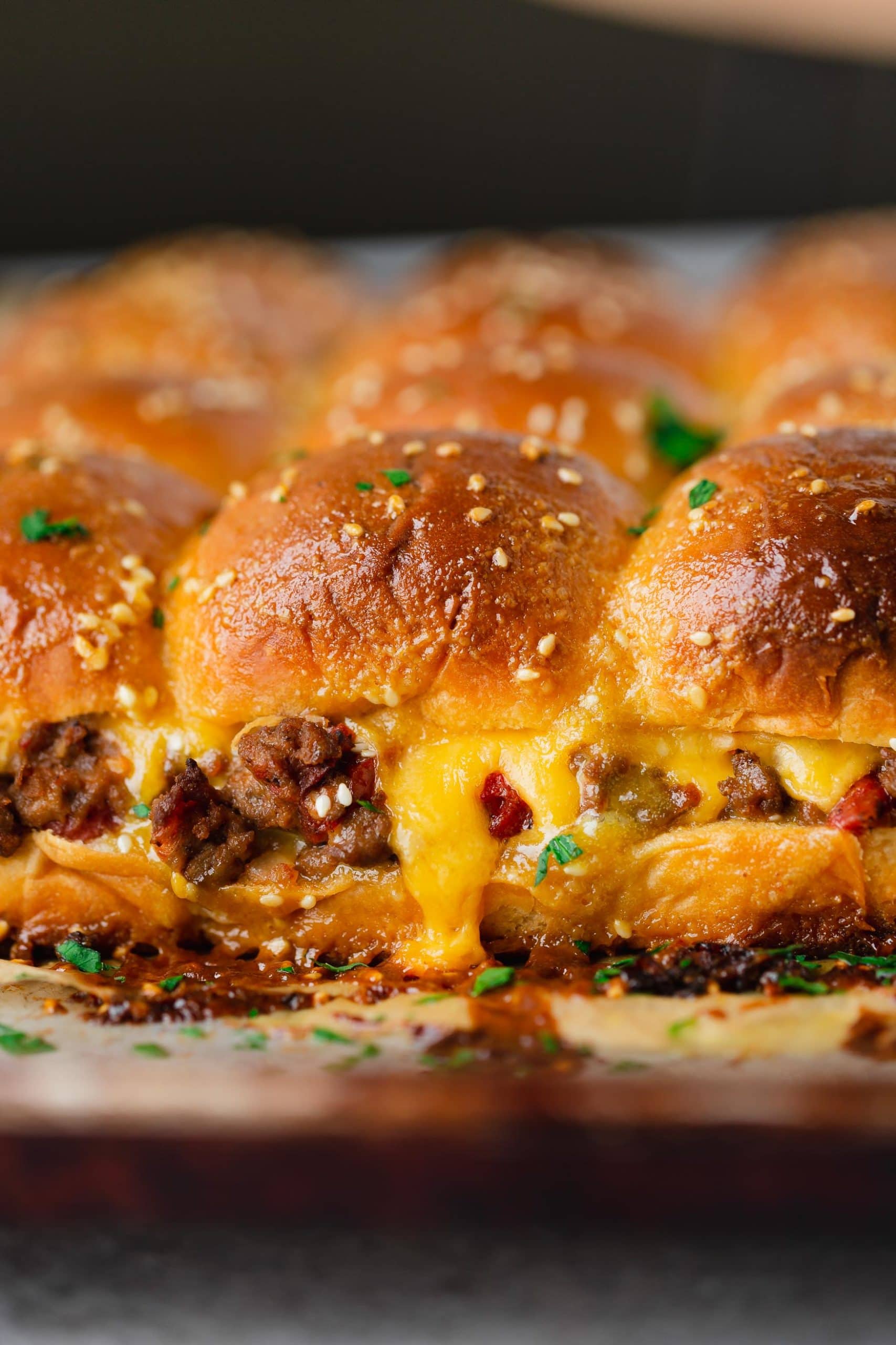 A panful of cheeseburger sliders. There is ground beef, chopped tomatoes and melted cheese on the buns and the buns are topped with sesame seeds and sauce.