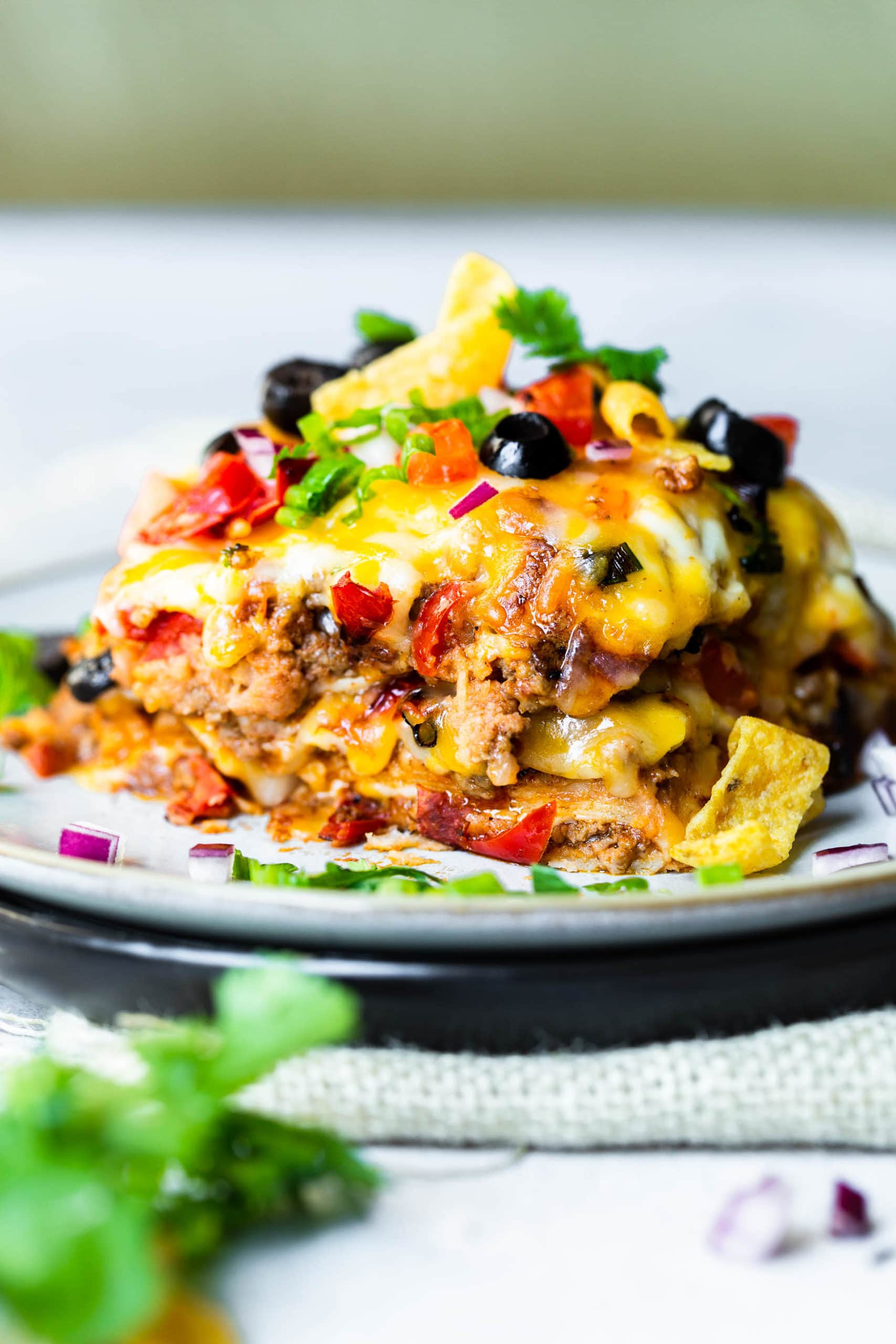 a photo of a piece of taco casserole on a saucer piled high with layers of cheesy ground beef, melted cheese on top, black beans, diced tomatoes and fresh cilantro.