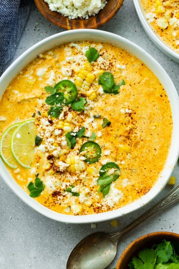 a photo of a large bowl of Mexican street corn soup topped with sliced jalapenos, chopped cilantro and lime wedges.