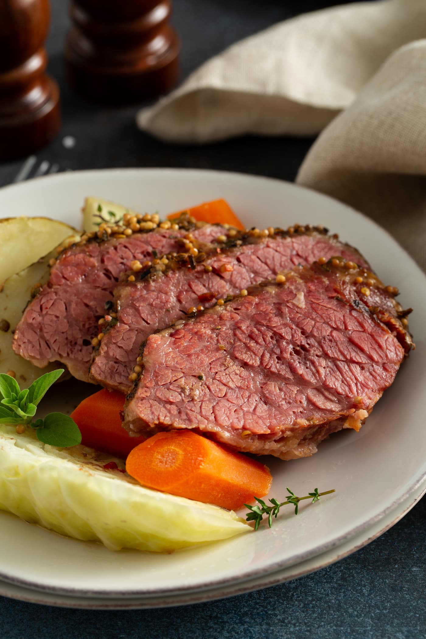 Three slices of corned beef next to carrots and potatoes and cabbage, all on a white serving plate. There are sprigs of green herbs on top.