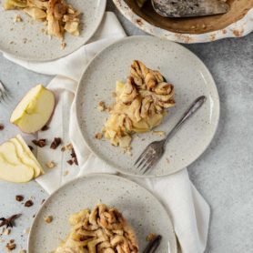 Slices of cinnamon roll apple pie on dessert plates. There are swirled cinnamon rolls as the crust and sliced apples in the background.