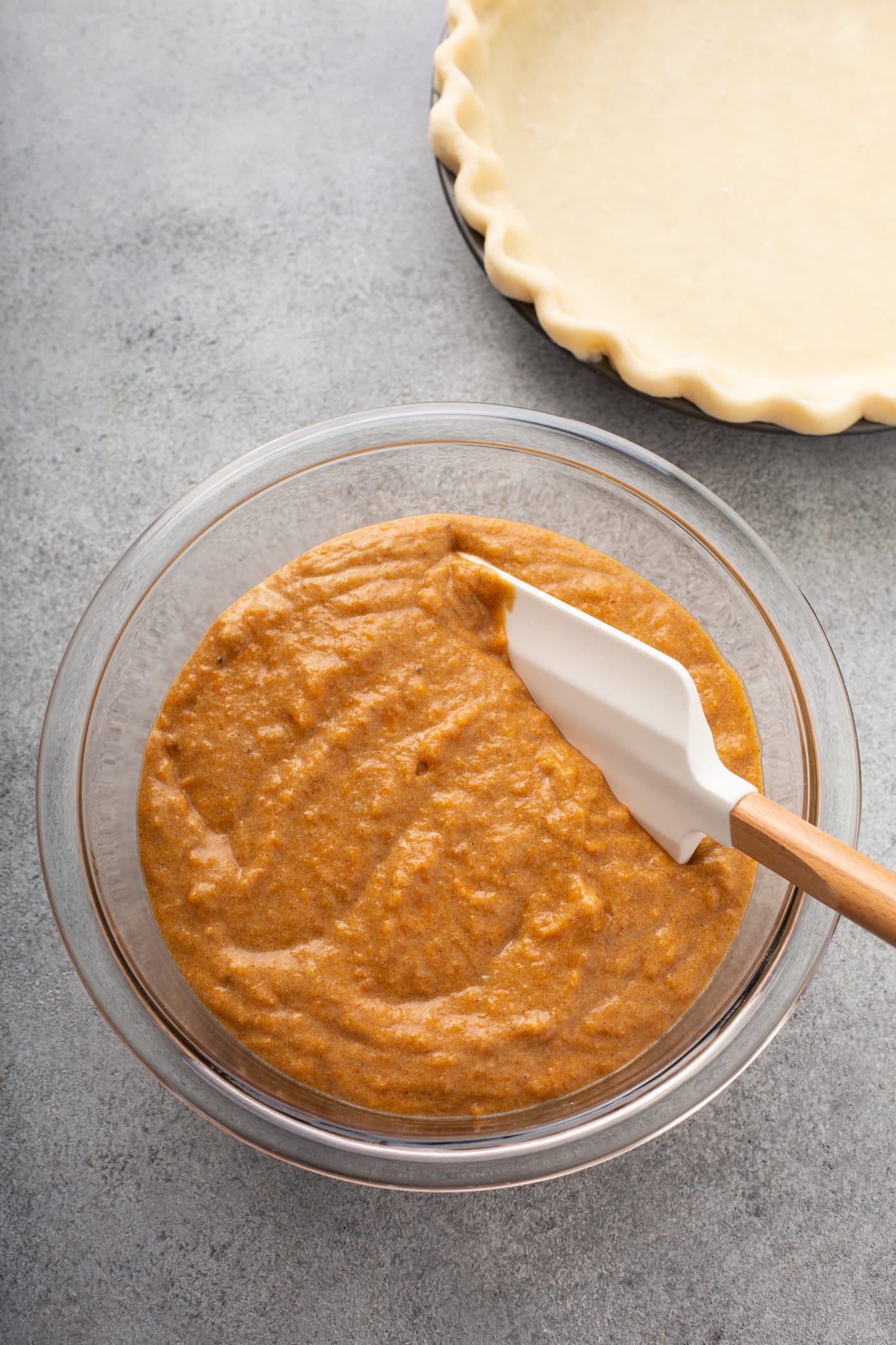 A photo of a glass bowl full of sweet potato pie filling with a rubber spatula in it. The bowl is sitting next to an empty pie crust.