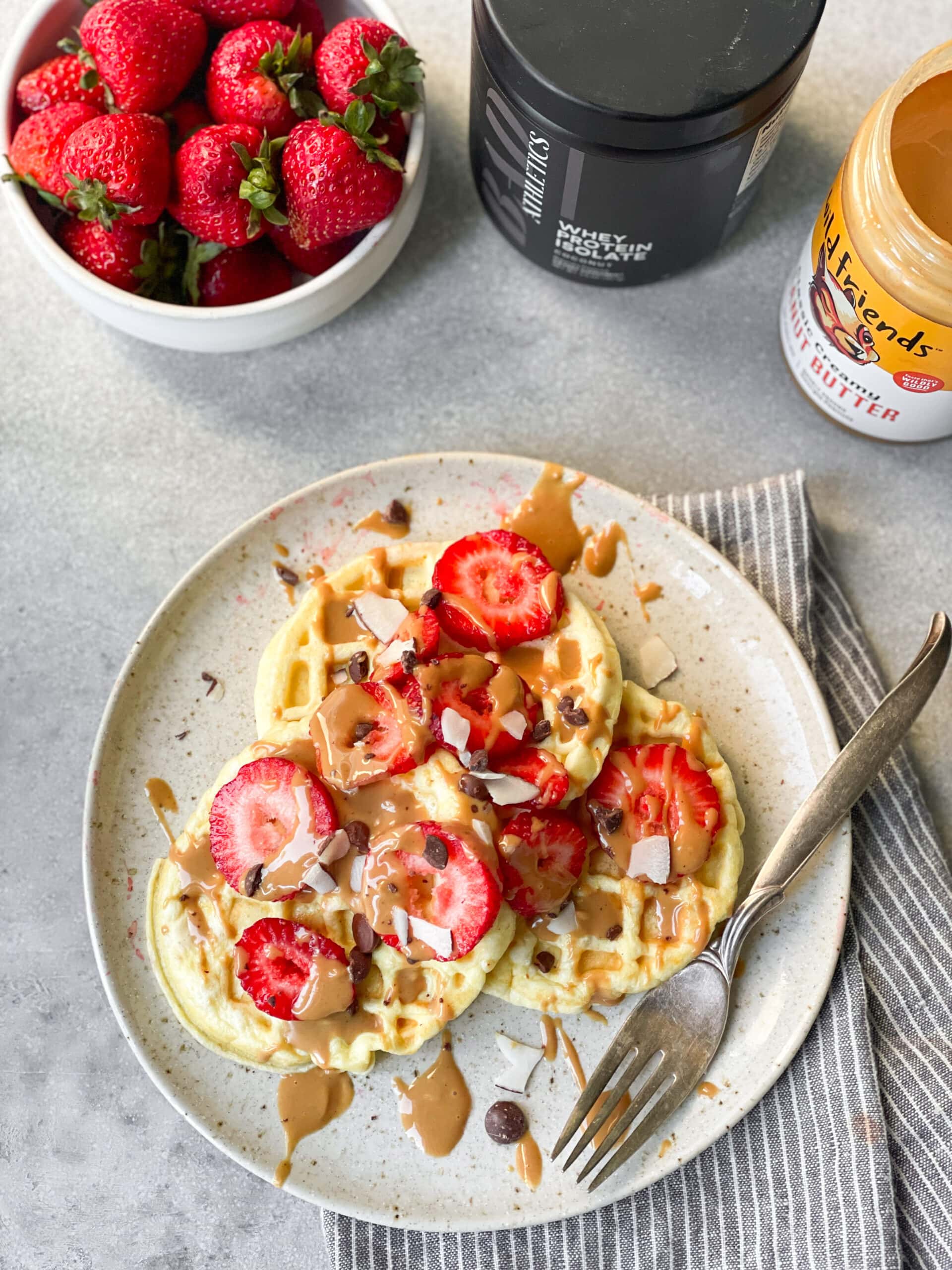 a photo of three small circular waffles topped with sliced strawberries, peanut butter drizzles, and chocolate chips on a white plate with a fork sitting on the side of the plate