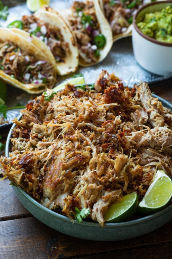 a photo of a bowl of shredded pork carnitas with three tacos in the background