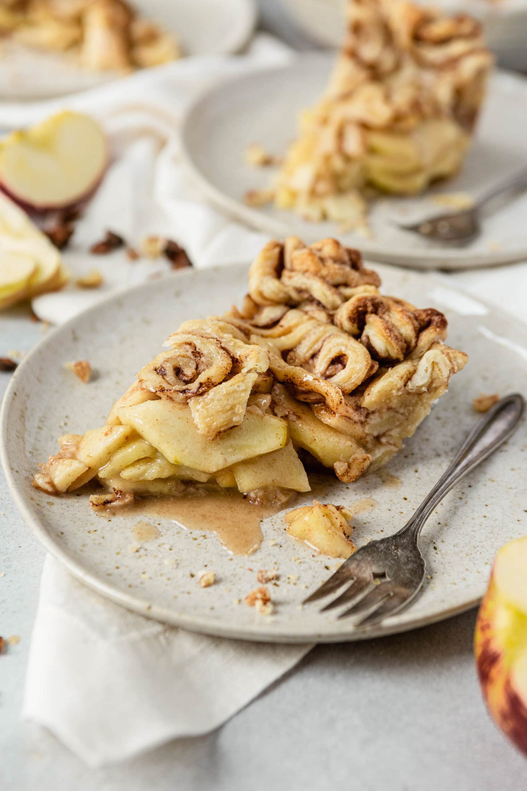 A slice of cinnamon roll apple pie with a fork next to it. There is another slice of pie on a plate in the background with slices of apples.