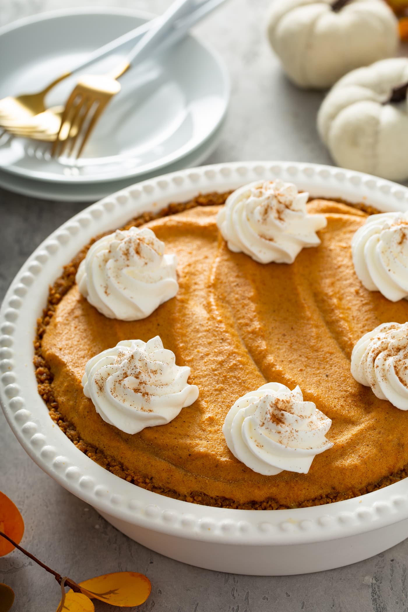 A whole pumpkin ice cream pie in a white pie dish. The pie is topped with dollops of whipped cream sprinkled with cinnamon. A stack of white dessert plates and two tiny white pumpkins are behind the pie.