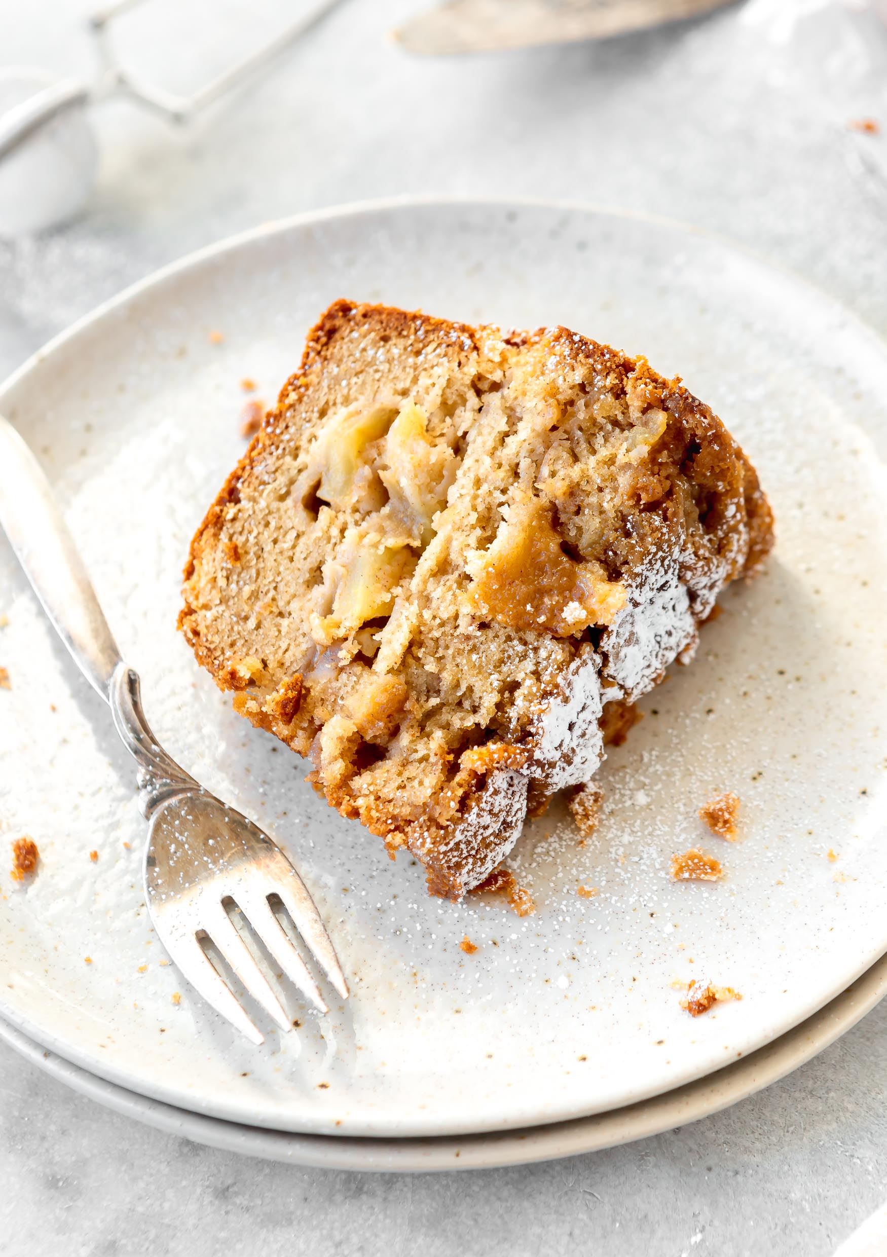 A slice of Jewish apple cake on a white dessert plate with a fork next to it. The cake is dense, and filled with apples. There is powdered sugar on the cake top.