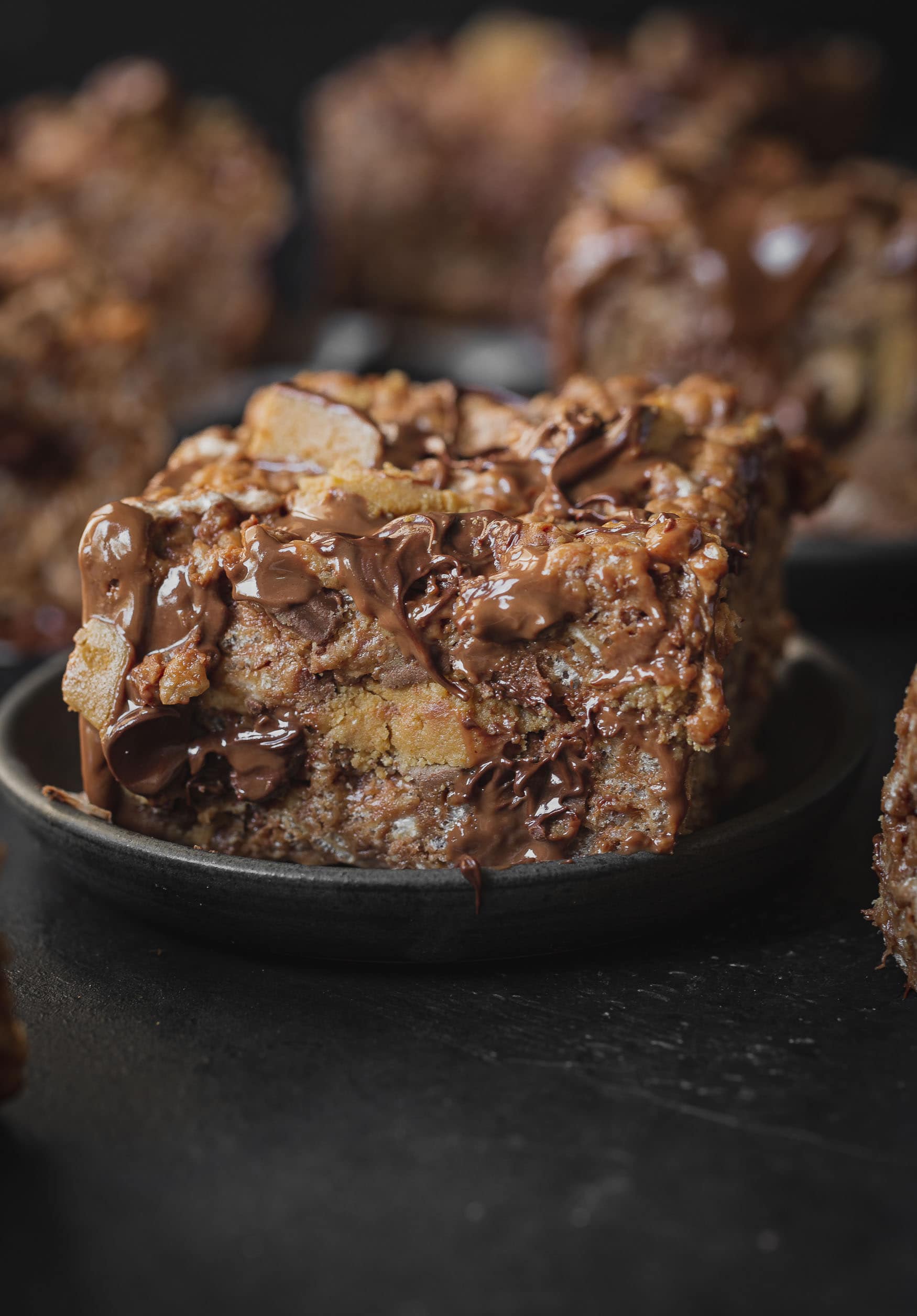 a photo of a single peanut butter rice krispies treats loaded with reese's peanut butter cups and melted chocolate chips on a dark gray plate with several more rice krispies treats in the background