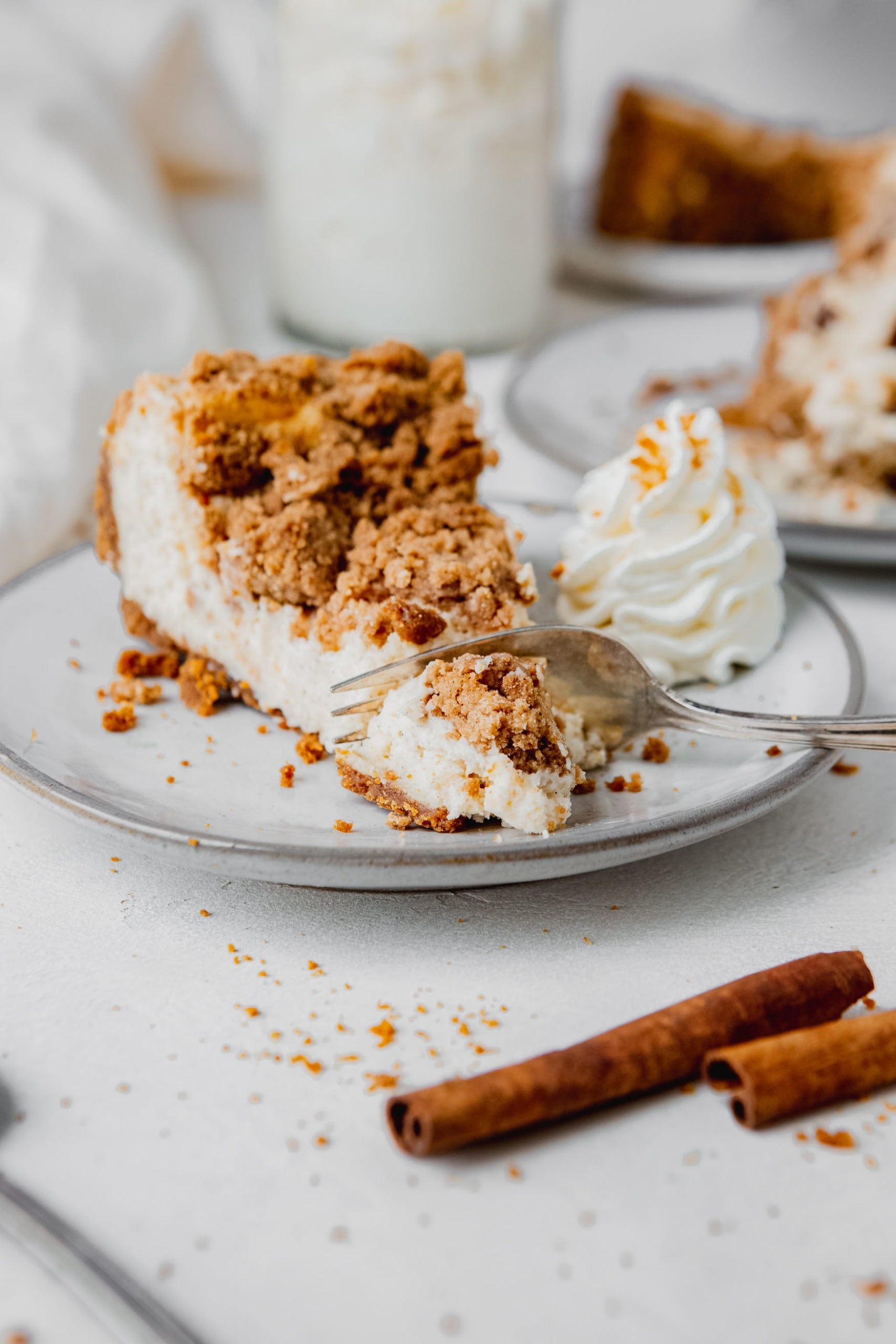 A slice of biscoff snickerdoodle cheesecake on a white dessert plate. There is a dollop of whipped cream on the plate next to the cheesecake. The creamy cheesecake has a cinnamon sugar snickerdoodle topping.