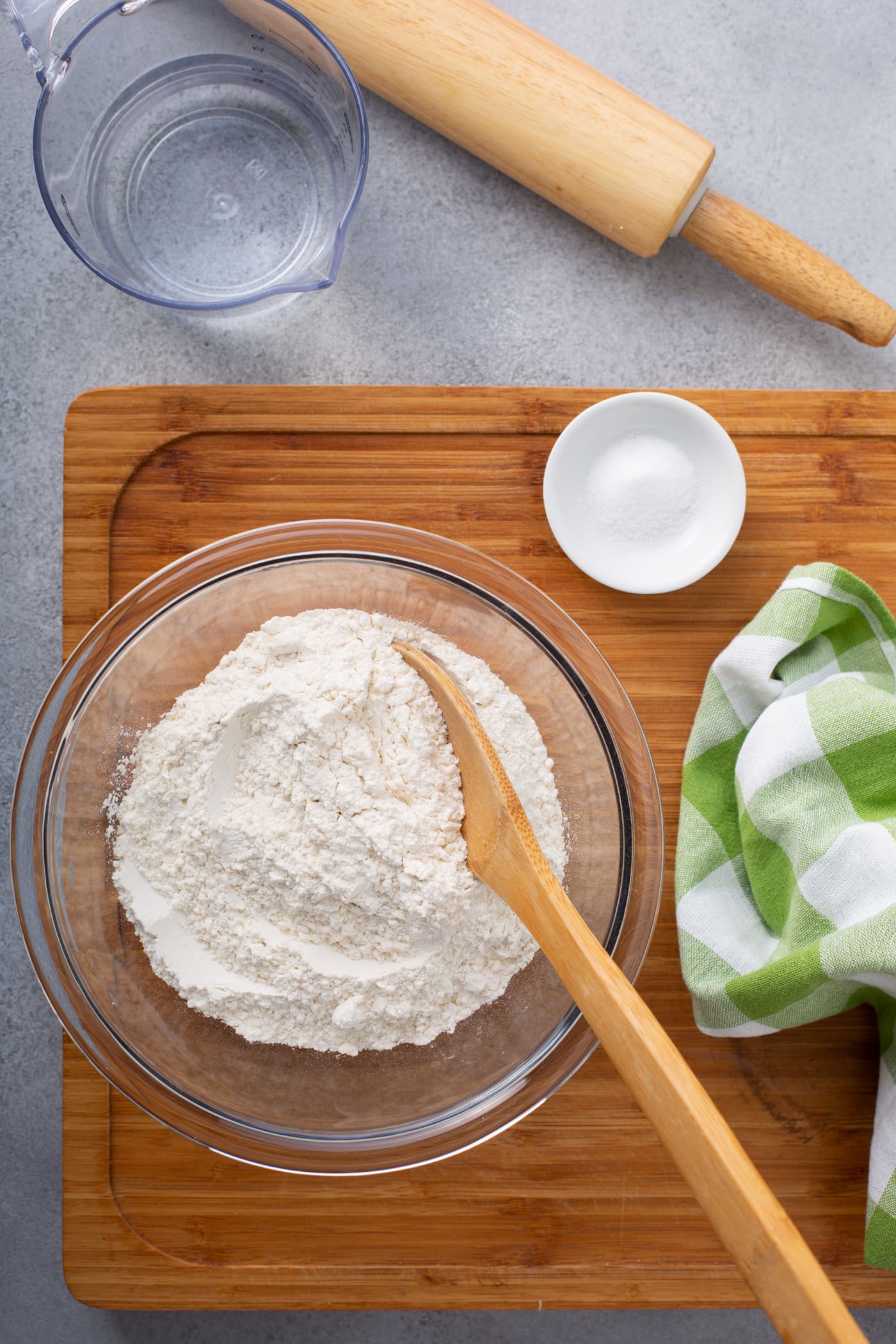 A photo of a glass bowl of flour with a wooden spoon in it and a small white bowl of salt sitting on a wooden cutting board with a glass measuring cup of water and wooden rolling pin on the side..