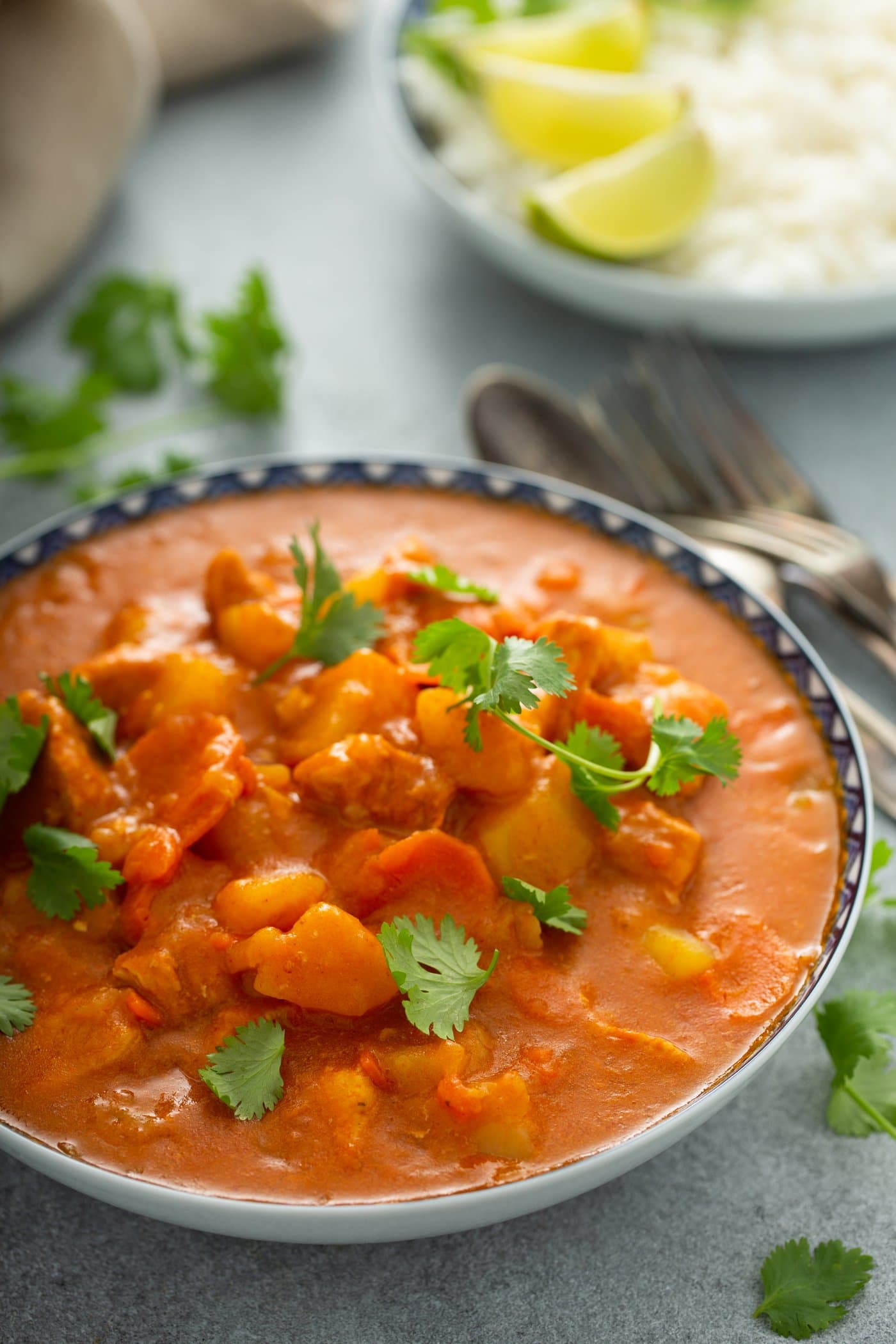 A bowl of chicken curry with chunks of chicken and potatoes and carrots in a curry sauce. Parsley leaves are scattered on top and on the table next to the curry. A fork and spoon and a plate with lime wedges are in the background.