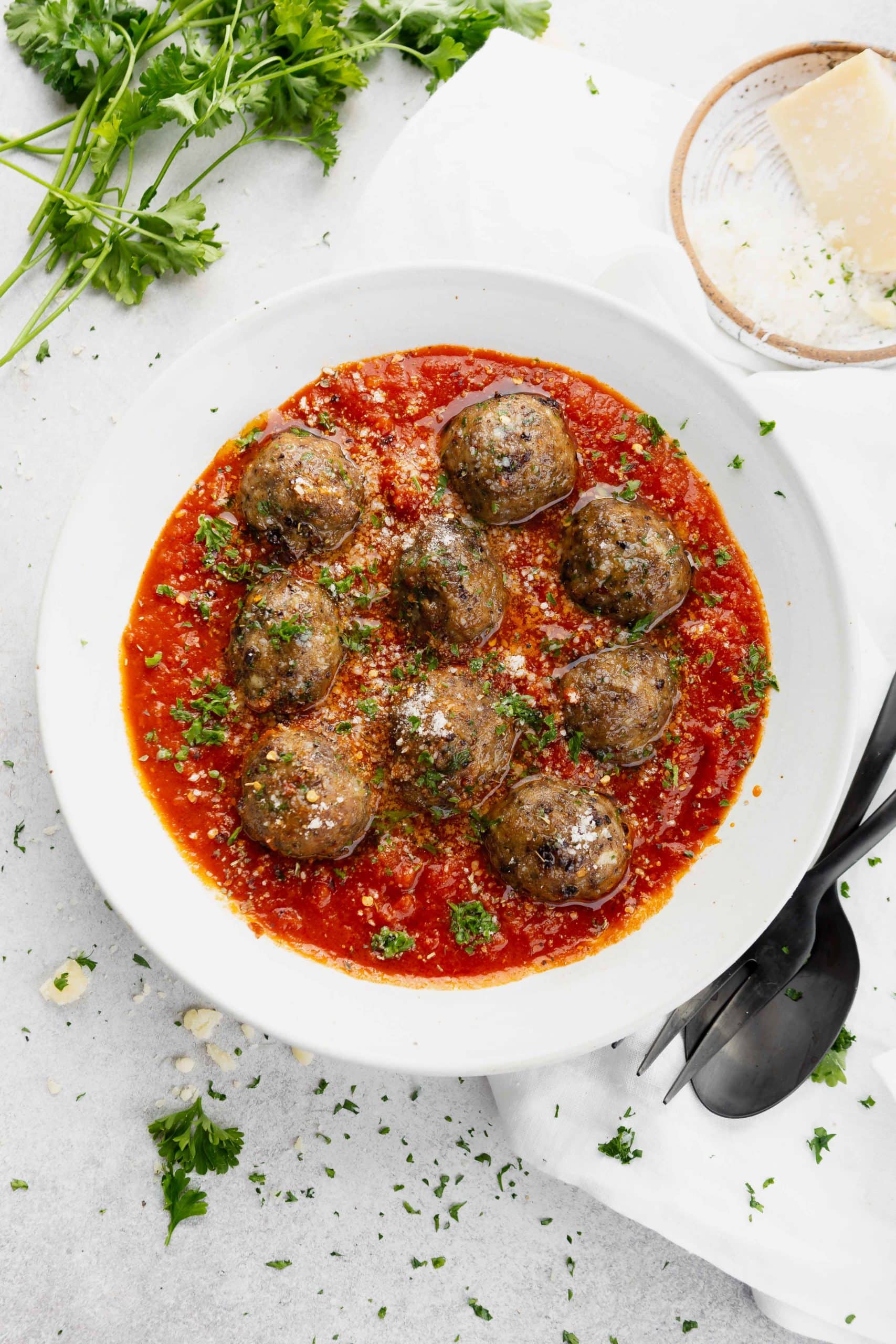 A bowl of meatballs in a tomato sauce. They are sprinkled with chopped parsley and there is a fork and spoon next to the bowl.