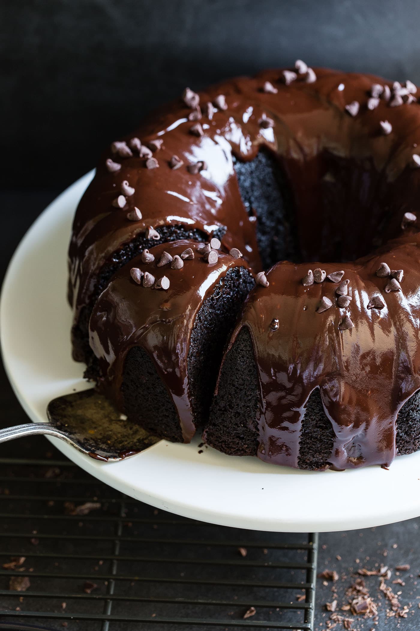 A chocolate zucchini bundt cake with chocolate glaze. The cake is on a white cake plate and a cake server is resting under a piece that has been cut.
