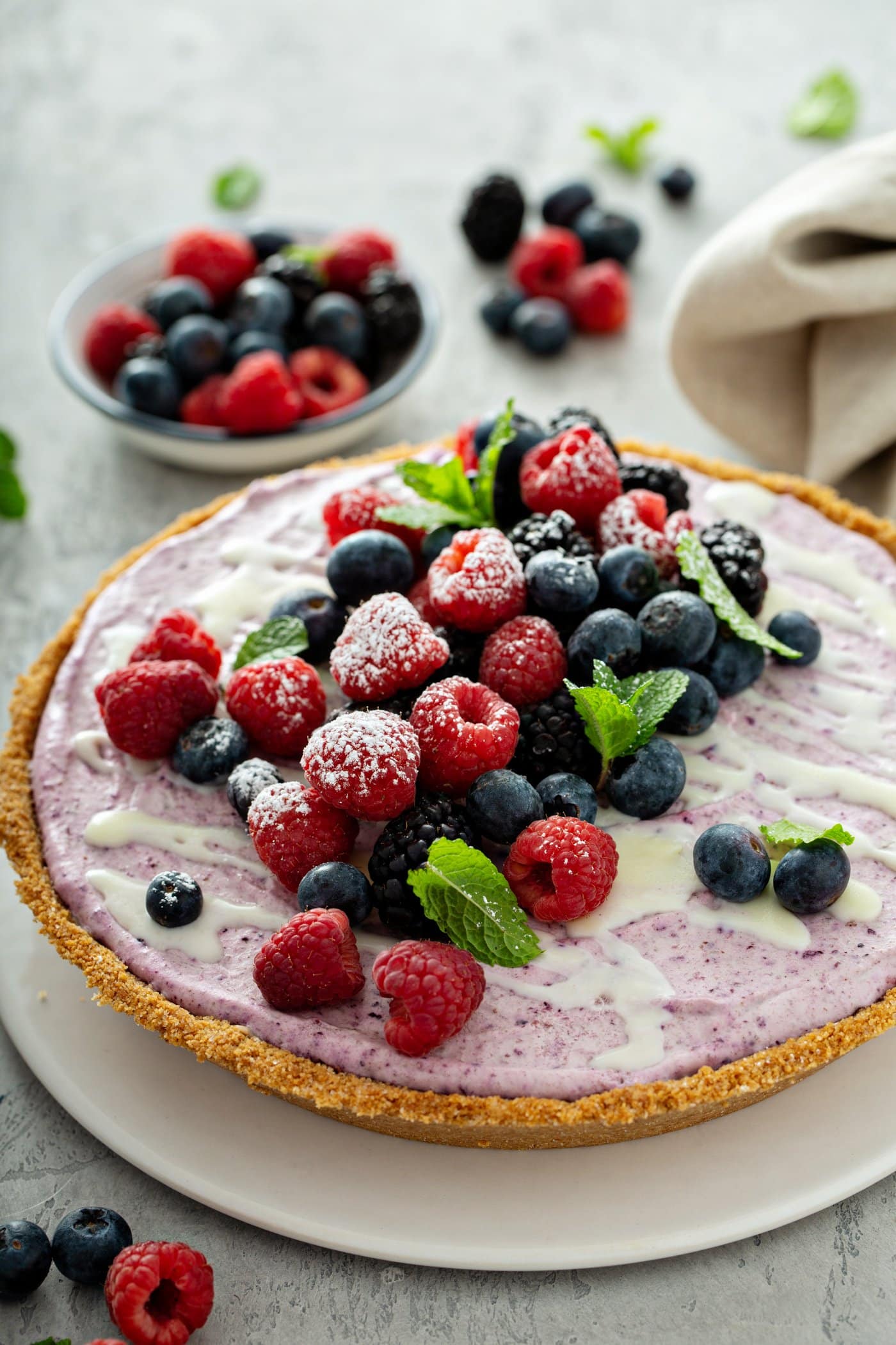 A whole white chocolate berry pie. The pie is topped with whole strawberries, raspberries, blue berries, blackberries, and mint leaves. There is a bowl of mixed berries in the background, and berries and a napkin on the table.
