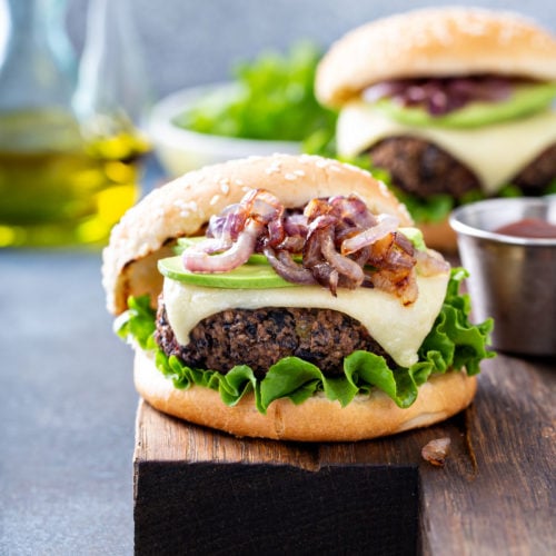 A table top with two black bean burgers on sesame seed buns. The burgers are topped with onions, avocados, cheese and lettuce. There is a bowl of lettuce and a container of oil in the background.