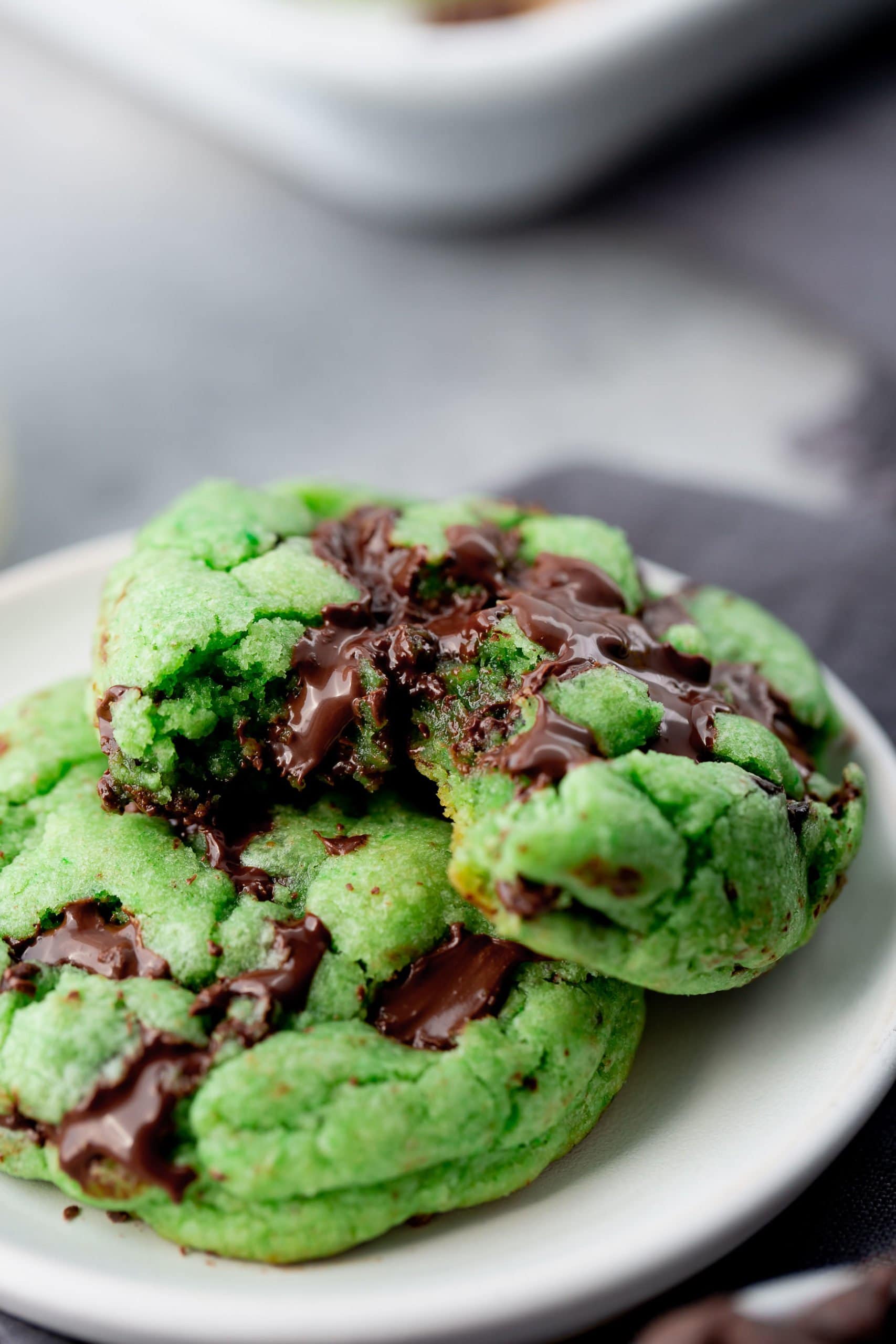 a photo of two green mint chocolate chip cookies on a white small plate. one cookie has a bite taken out of it.