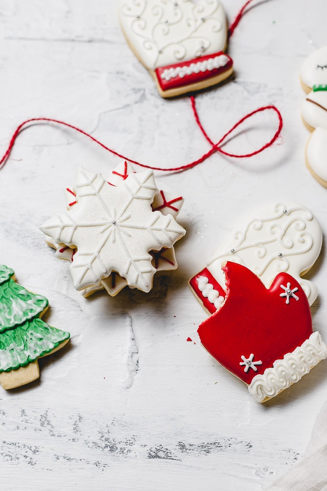 Christmas sugar cookies decorated with royal icing. There are white snowflakes, red mittens, white mittens with red trim, and a green tree with white snow.