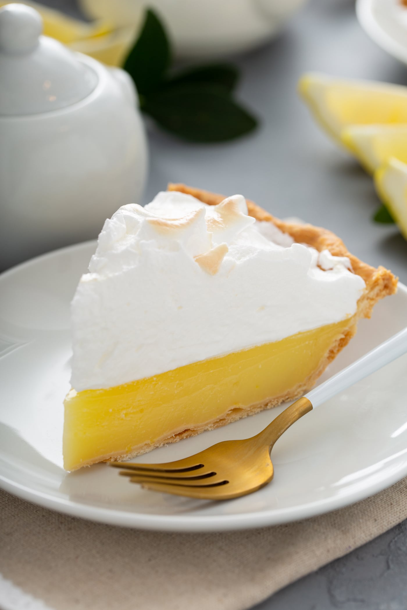 A slice of lemon meringue pie on a white dessert plate. A fork is next to the slice of pie. The filling is bright yellow and the meringue is lightly toasted.