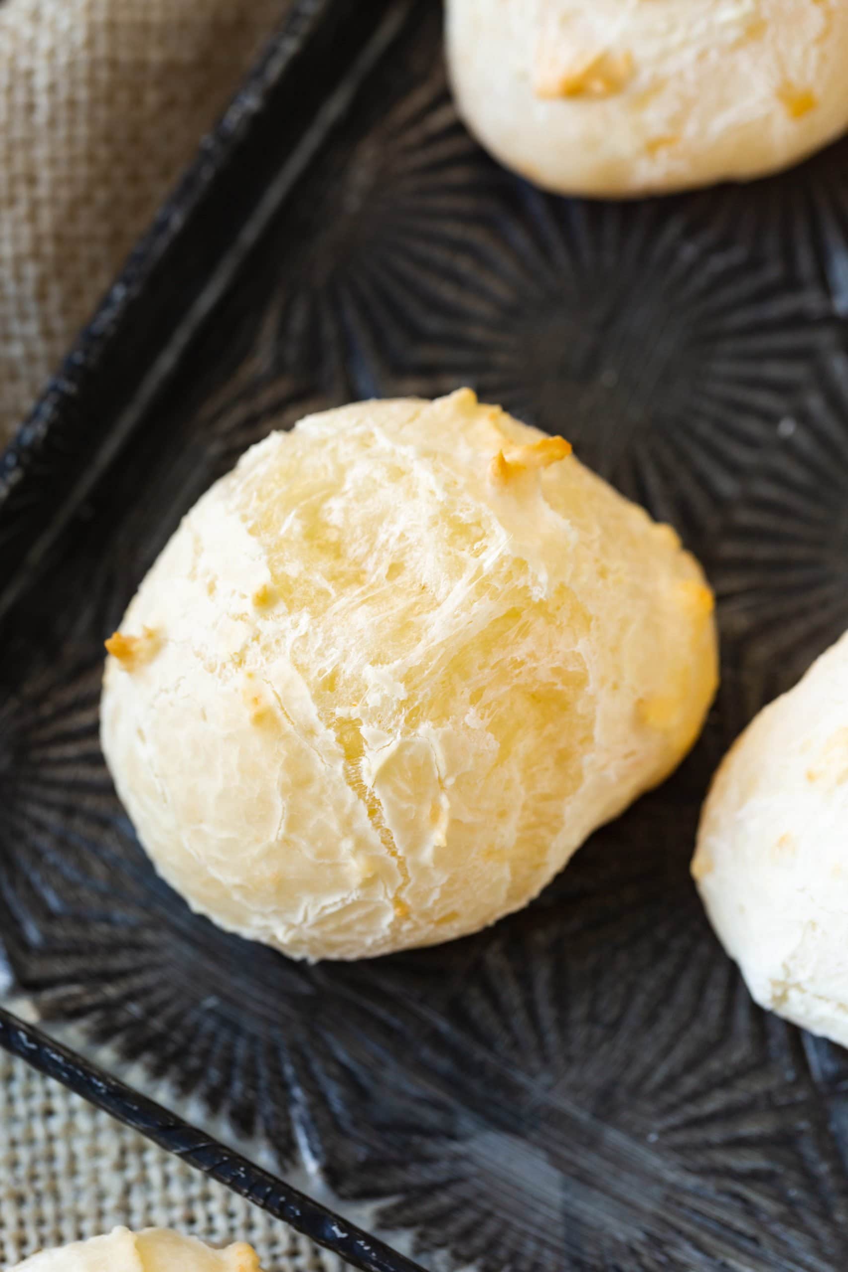a photo of a single piece of pao de queijo sitting on a baking sheet next to other pieces baked to golden perfection.