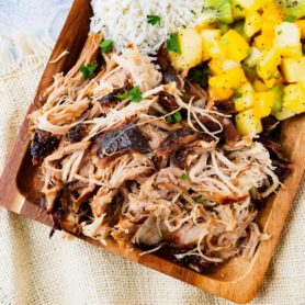 a photo of a rectangular wooden platter full of kalua pulled pork, herbed rice, tropical fruit salad and golden Hawaiian rolls.