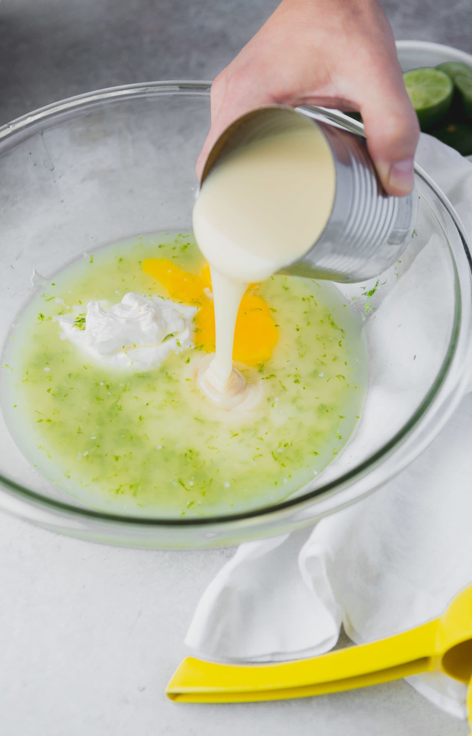 Cream being poured from a measuring cup into a mixing bowl with eggs, lime juice, and sour cream.