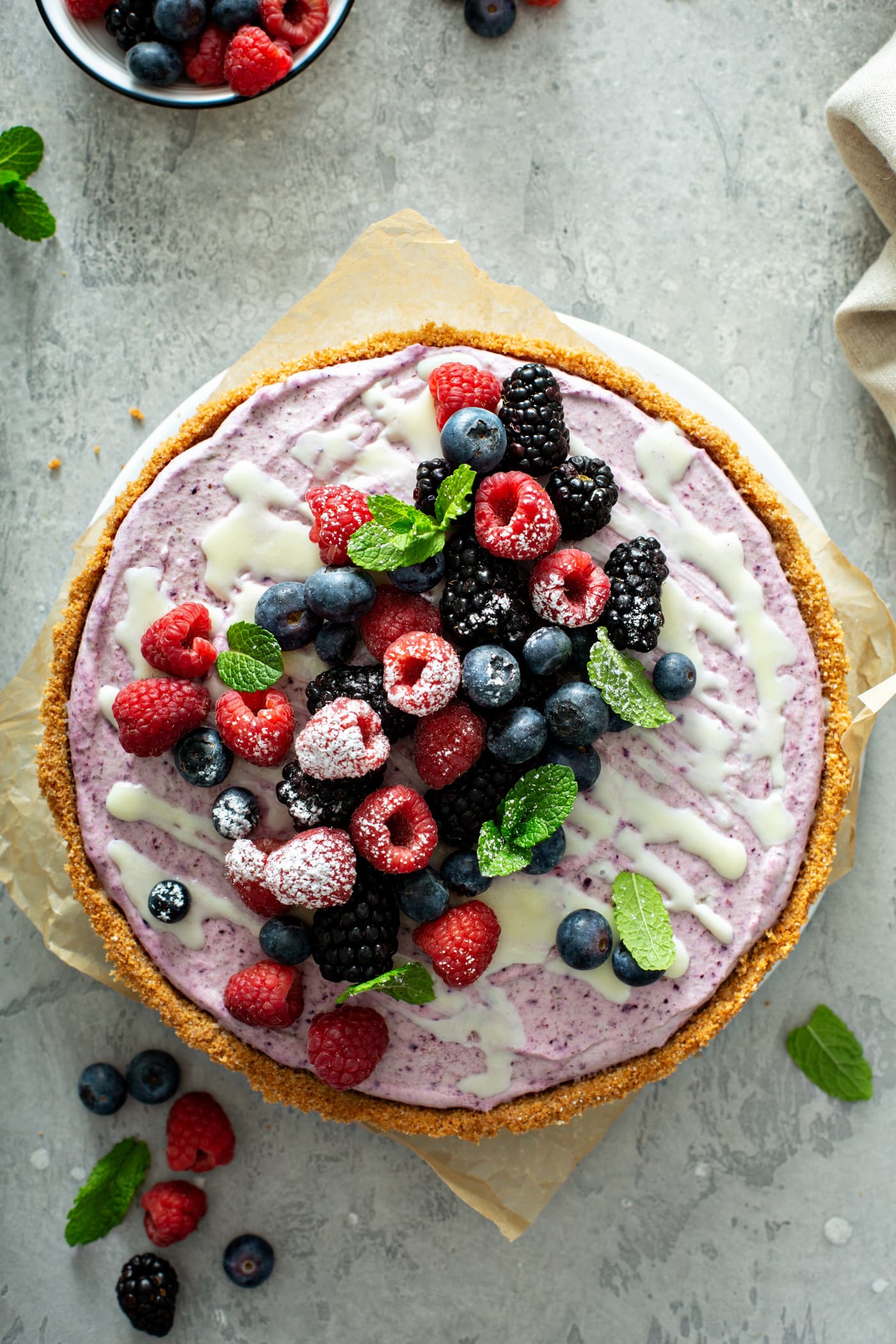 A whole white chocolate berry pie with mixed berries and mint leaves on top. A white chocolate sauce is drizzled over the top of the pie.