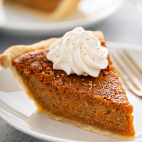 A slice of sweet potato pie with a dollop of whipped cream on top, sitting on a white plate with a fork beside it.