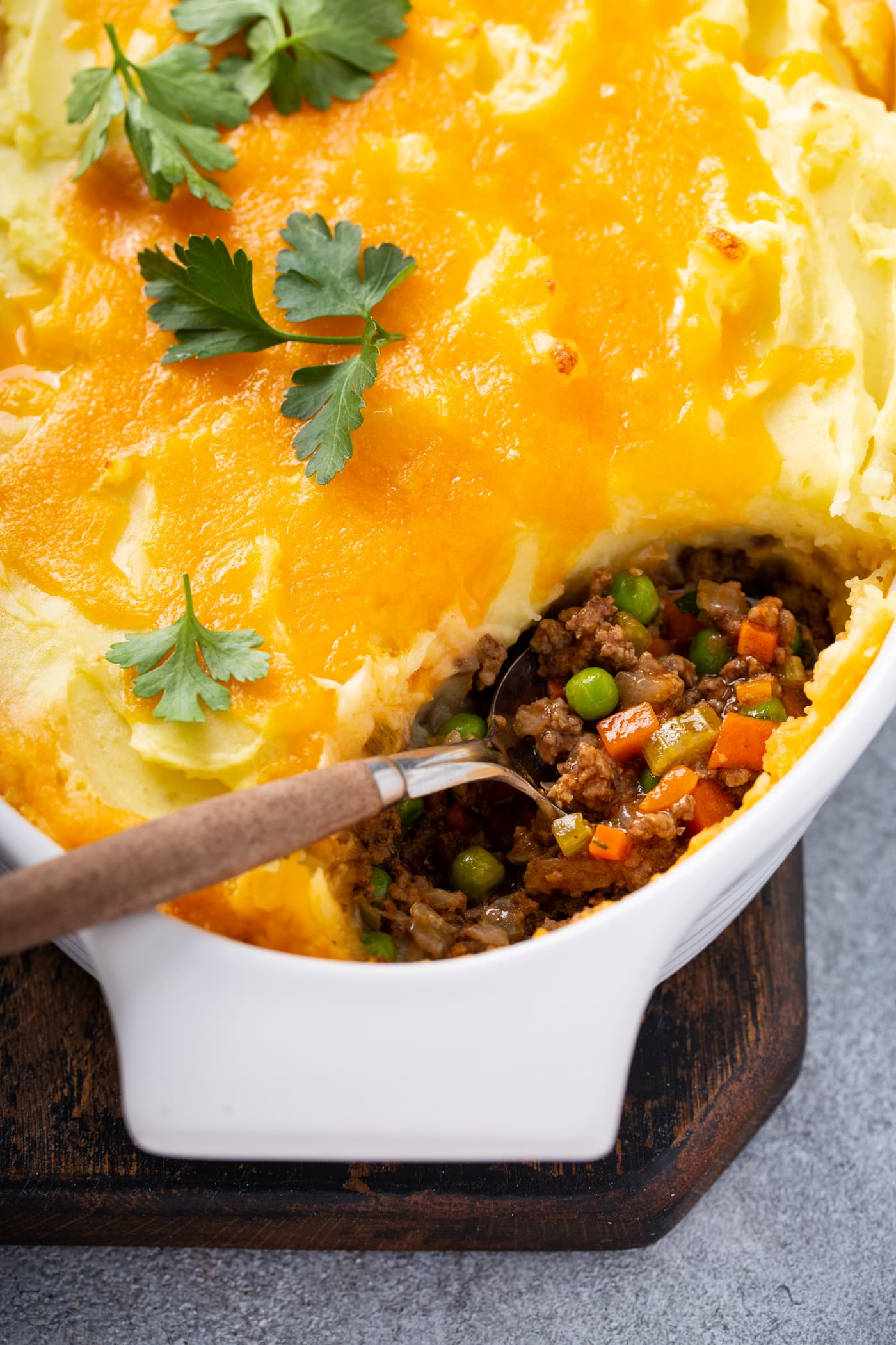 A casserole dish containing shepherd's pie with one spoonful removed and the serving spoon resting in the dish. You can see hamburger, carrots, and peas in the pie and it is topped with mashed potatoes, and melted cheddar cheese. There is a few parsley leaves on top.