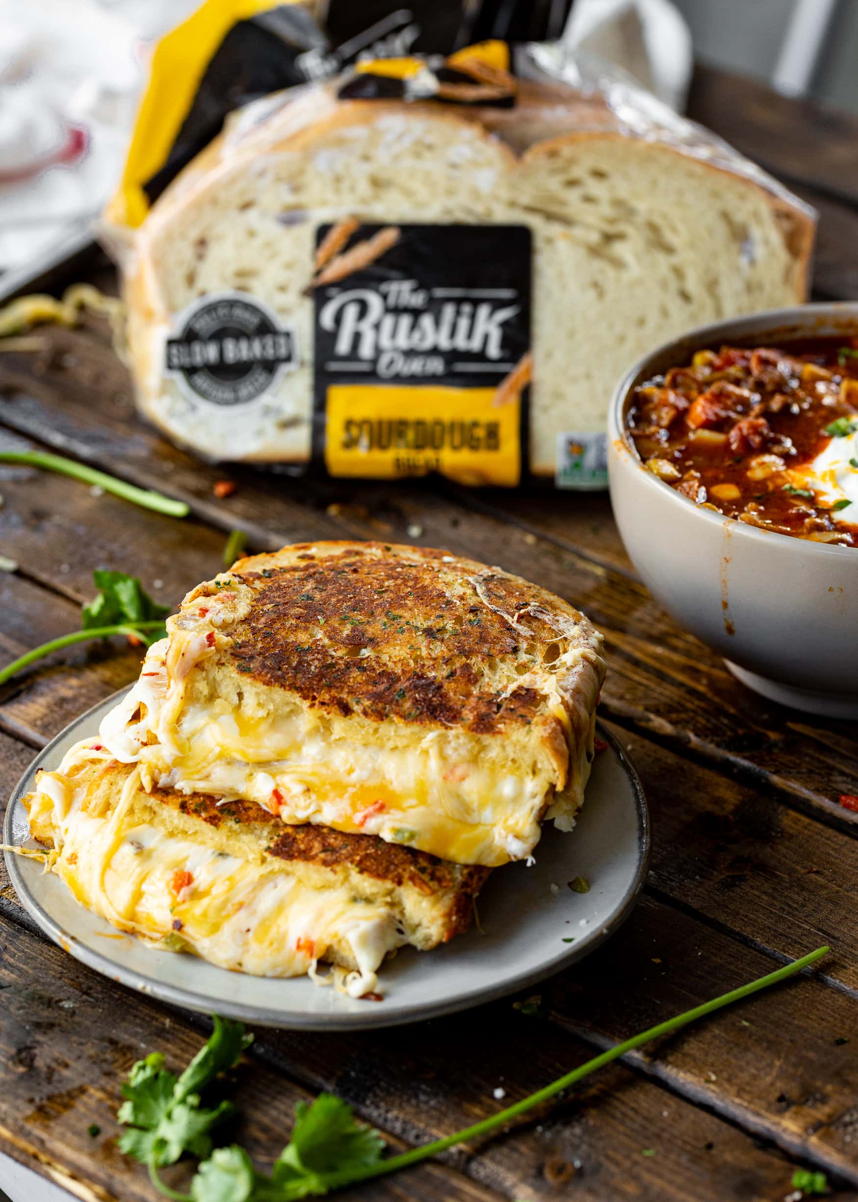 a photo of a grilled cheese sandwich that has been cut in half and stacked on top of each other sitting on a small plate with a bowl of taco stew and a bag of sourdough bread in the background.