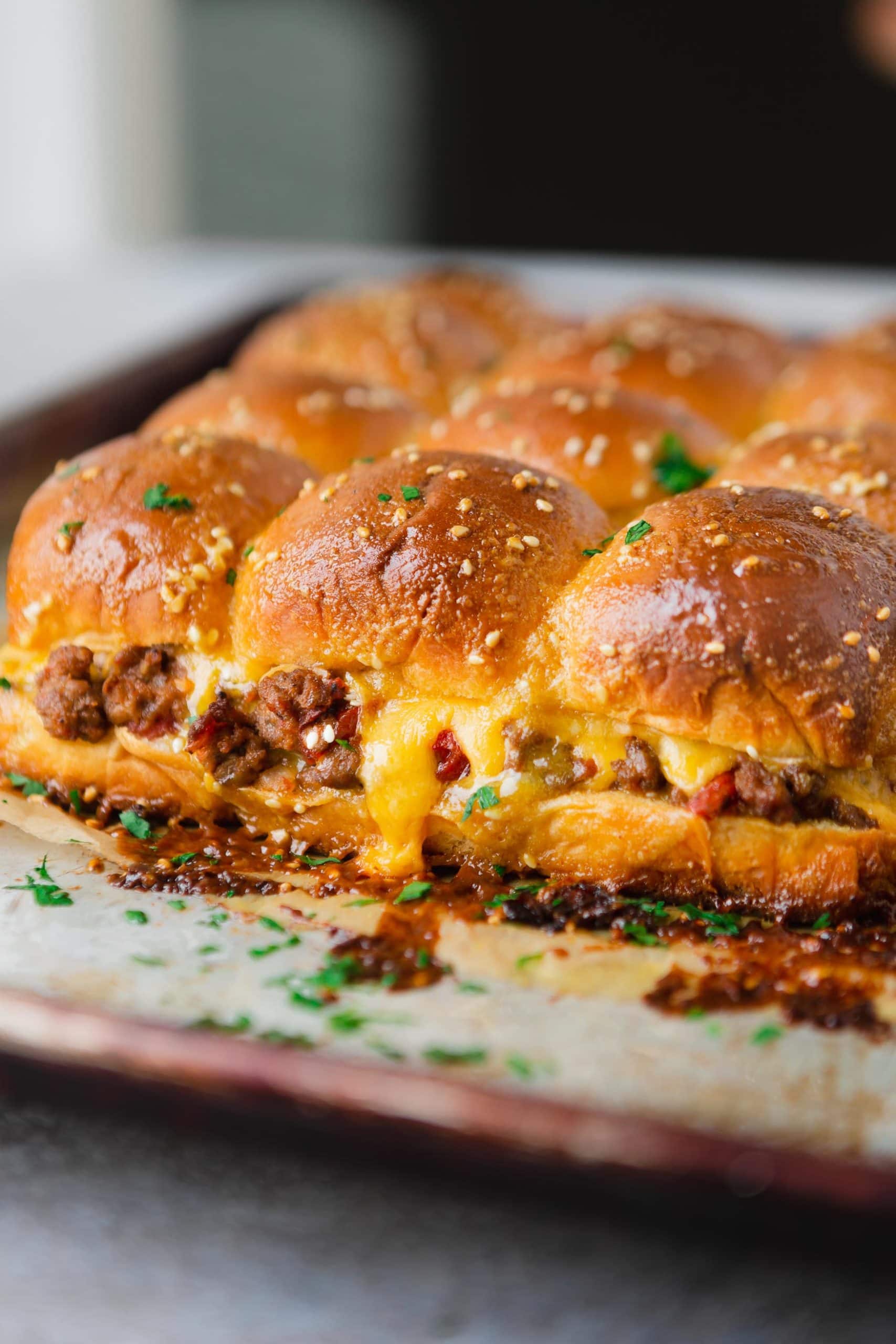 A panful of cheeseburger sliders. The cheese is oozing from the rolls and you can see ground beef and chunks of tomatoes.