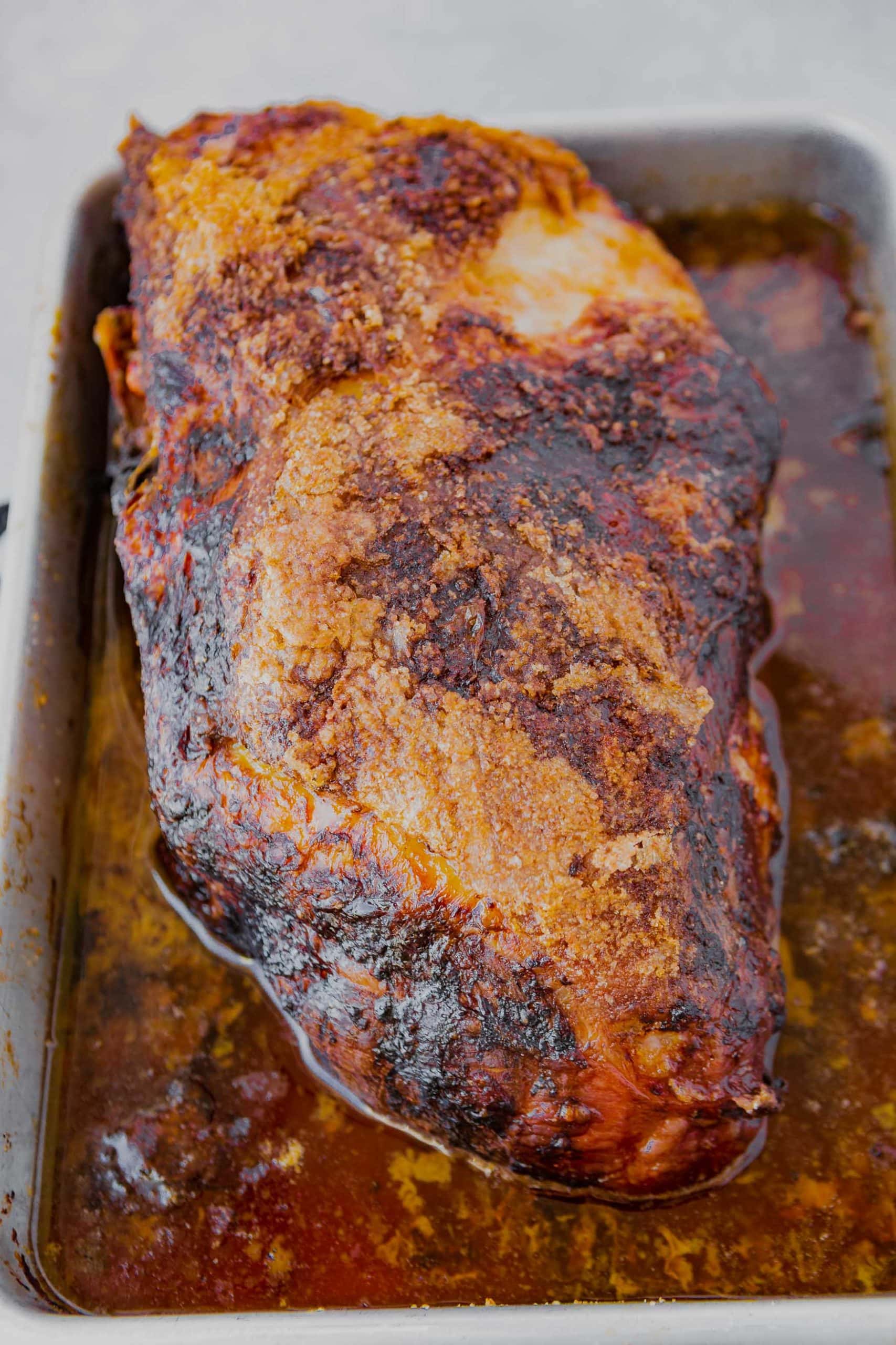 a perfectly roasted whole pork butt in a white baking dish sitting into its juices ready to be shredded