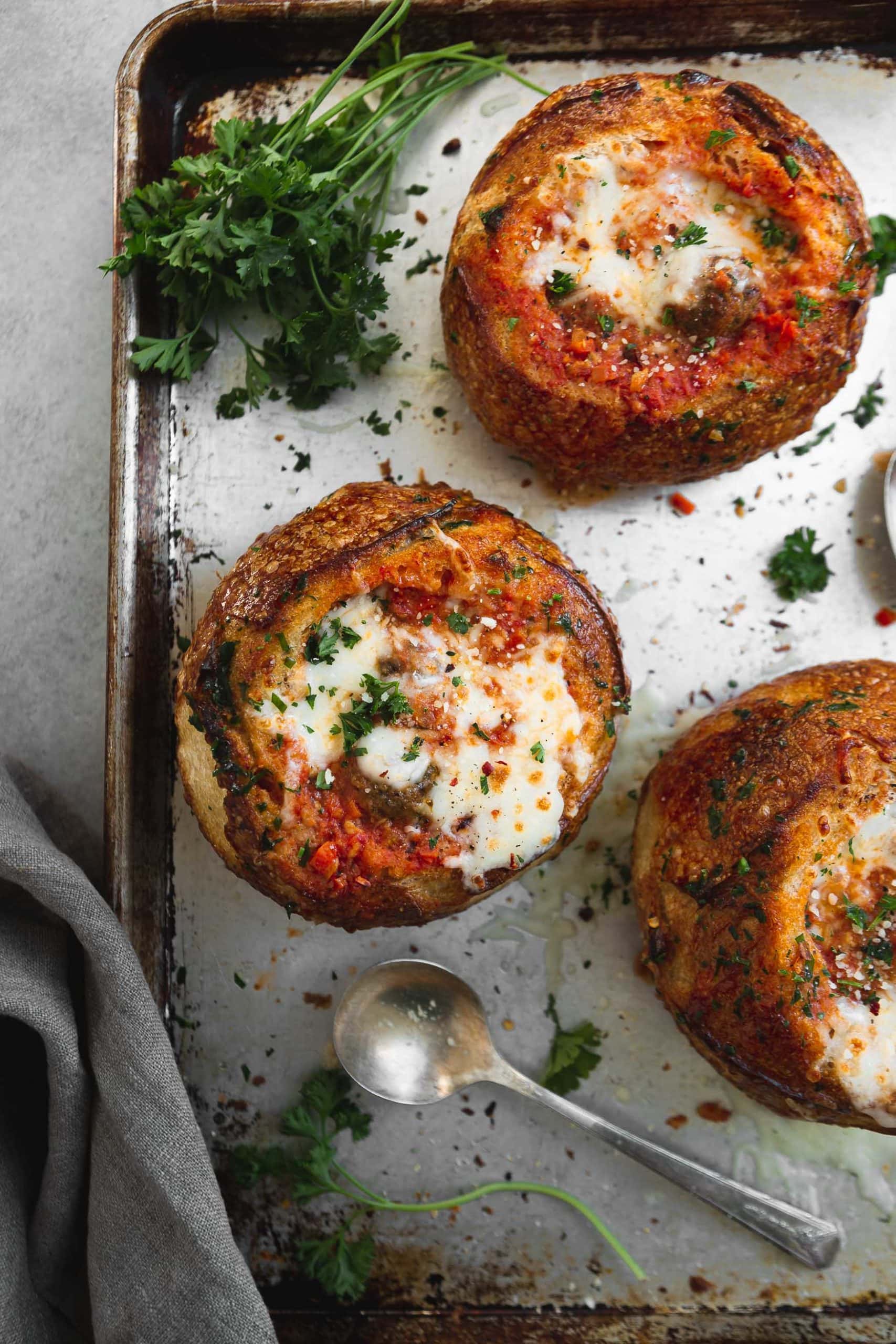 A baking sheet with three bread bowls filled with parmesan meatball soup. They have been baked to a golden brown and the cheese on top is melted to perfection. They are sprinkled with fresh parsley.