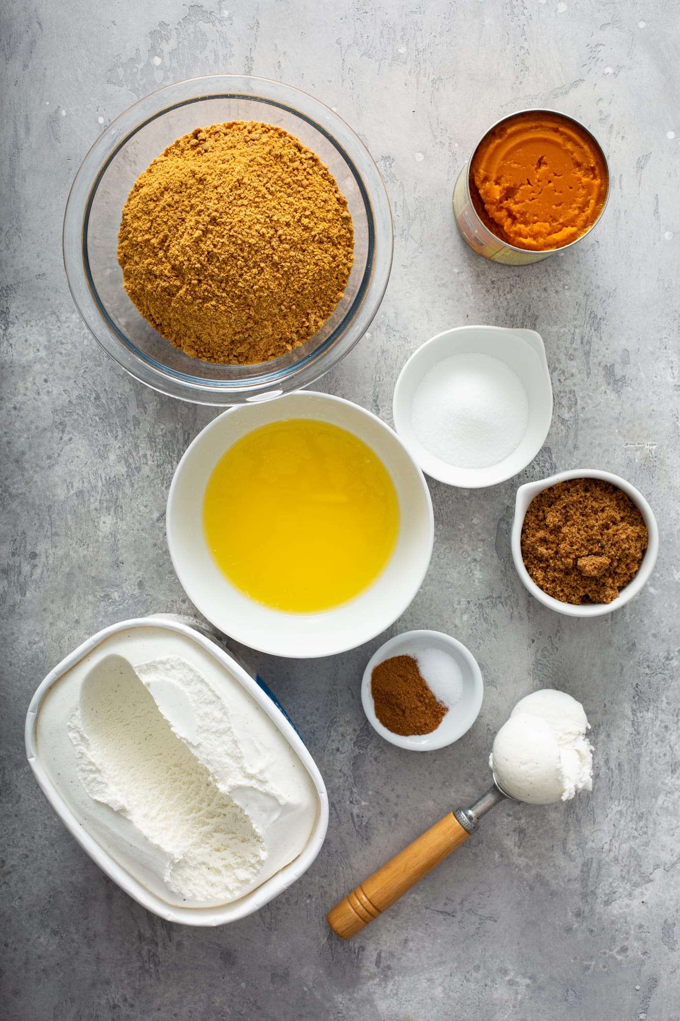 Small containers of ingredients for pumpkin ice cream pie. There are containers of pumpkin, gingersnap crumbs, butter, spices, sugar, brown sugar, and vanilla ice cream. 
