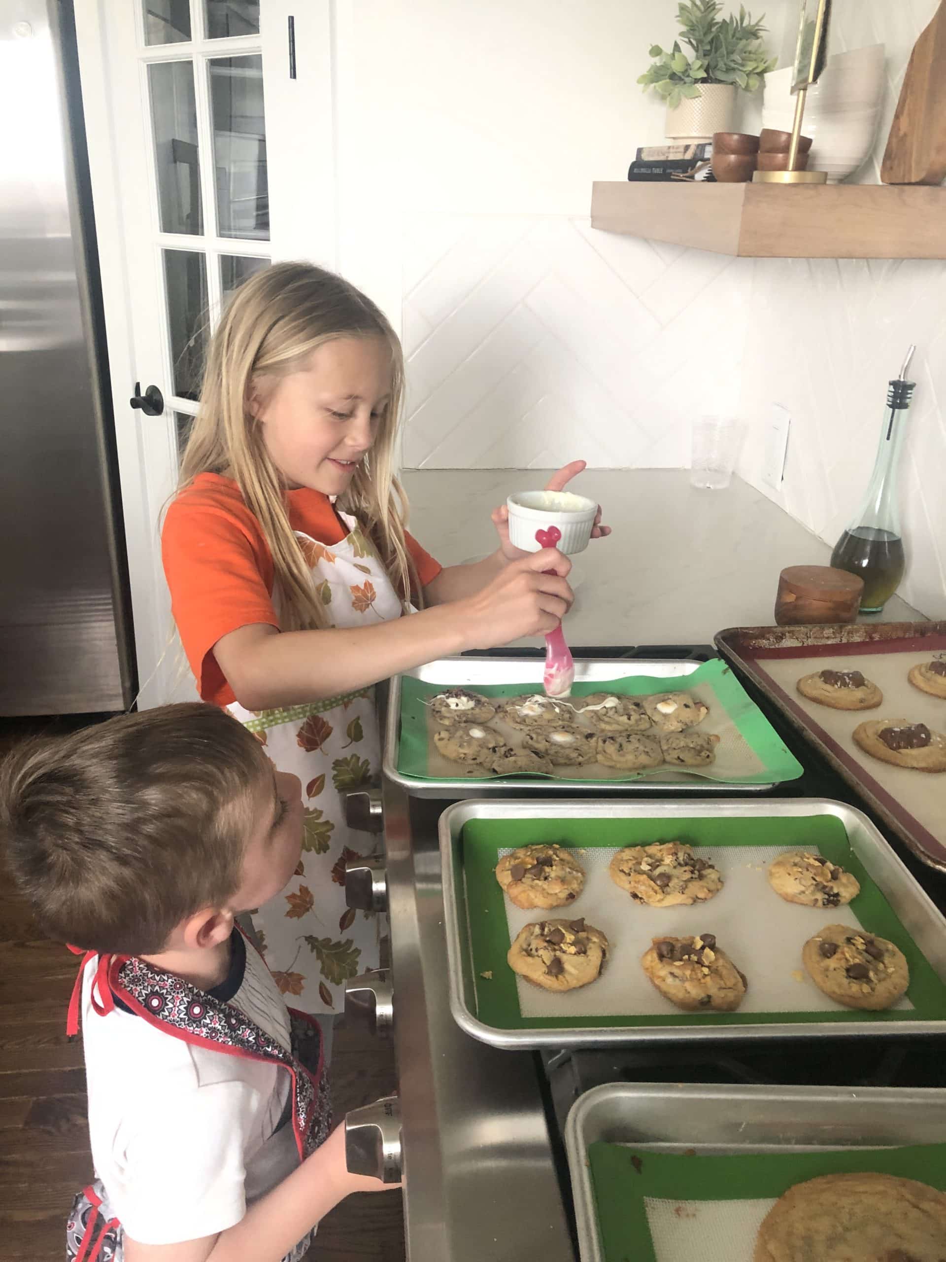 A photo of a young girl spooning melted white chocolate over chocolate chip cookies with her little brother watching.