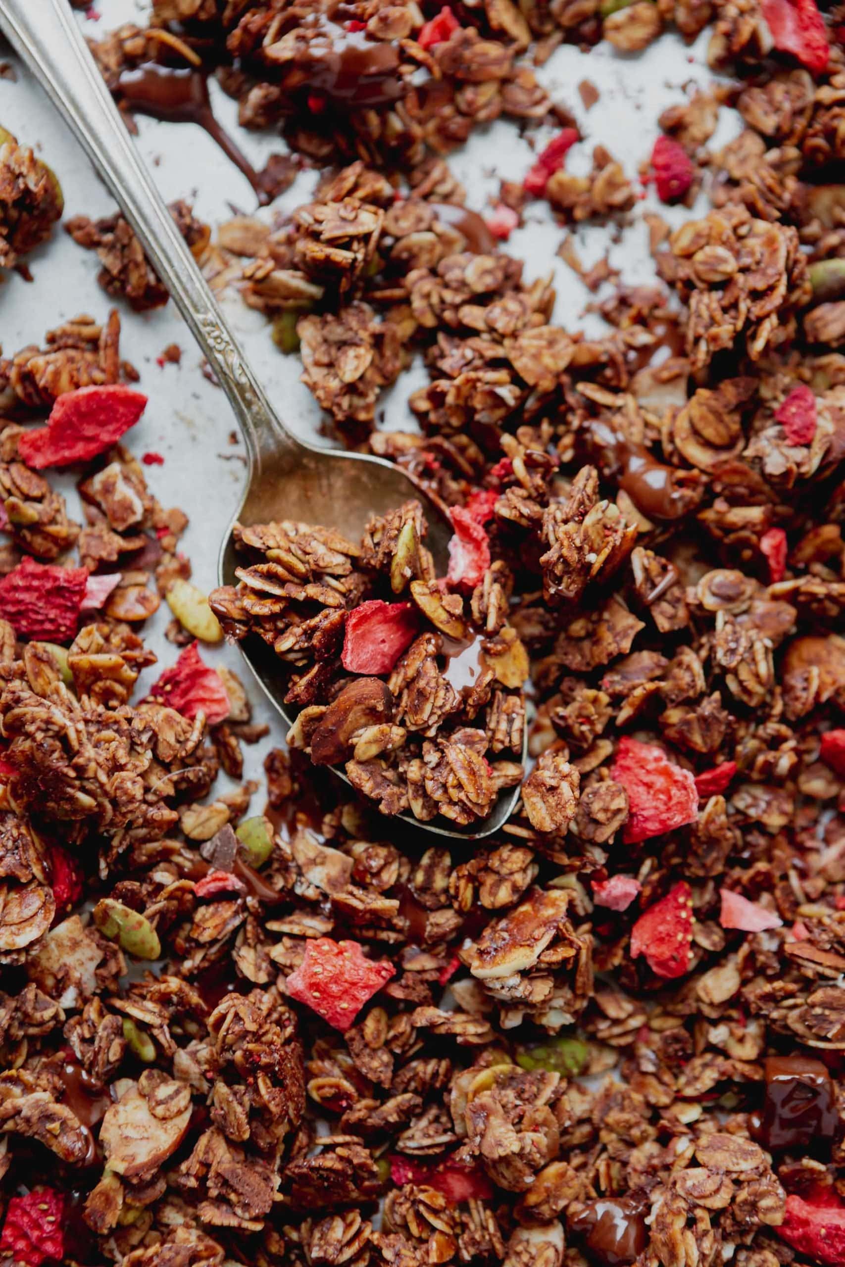 Chocolate granola with dehydrated strawberries on a baking pan with a spoon.