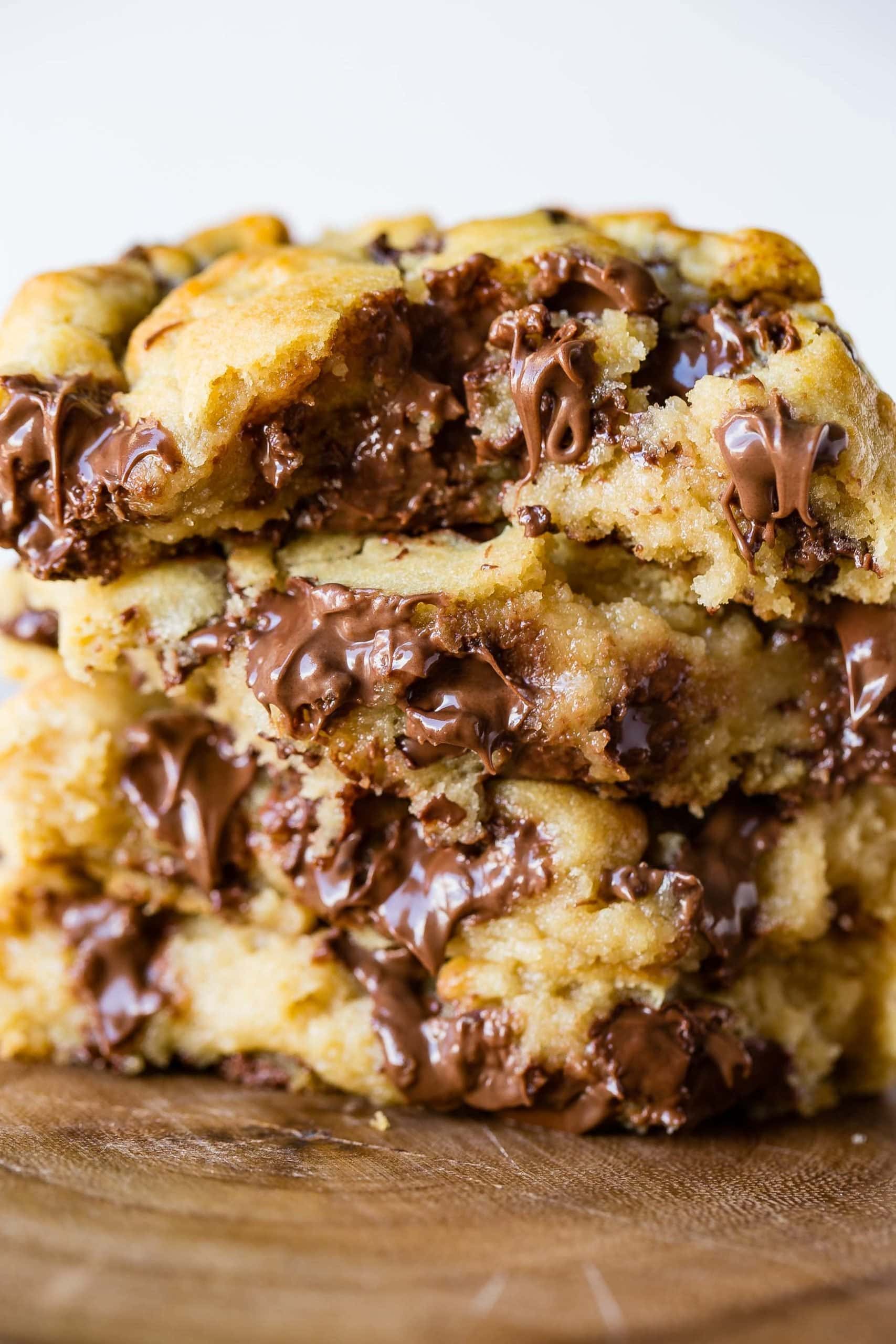 a closeup photo of four thick melty chocolate chip cookies that have been broken in half stacked on top of each other on top of a wooden plate.