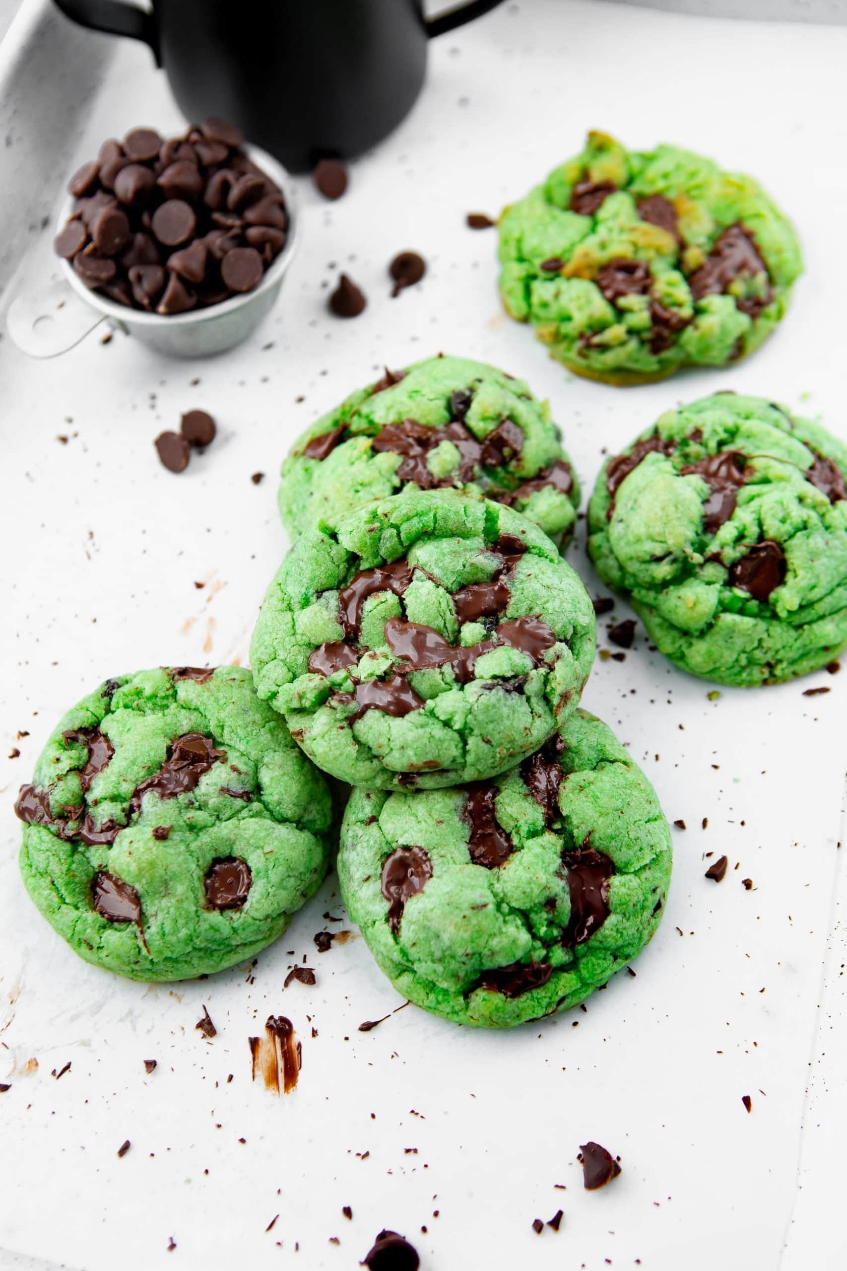 a photo of several green mint chocolate chip cookies on a parchment lined baking sheet with a cup of chocolate chips sitting next them.