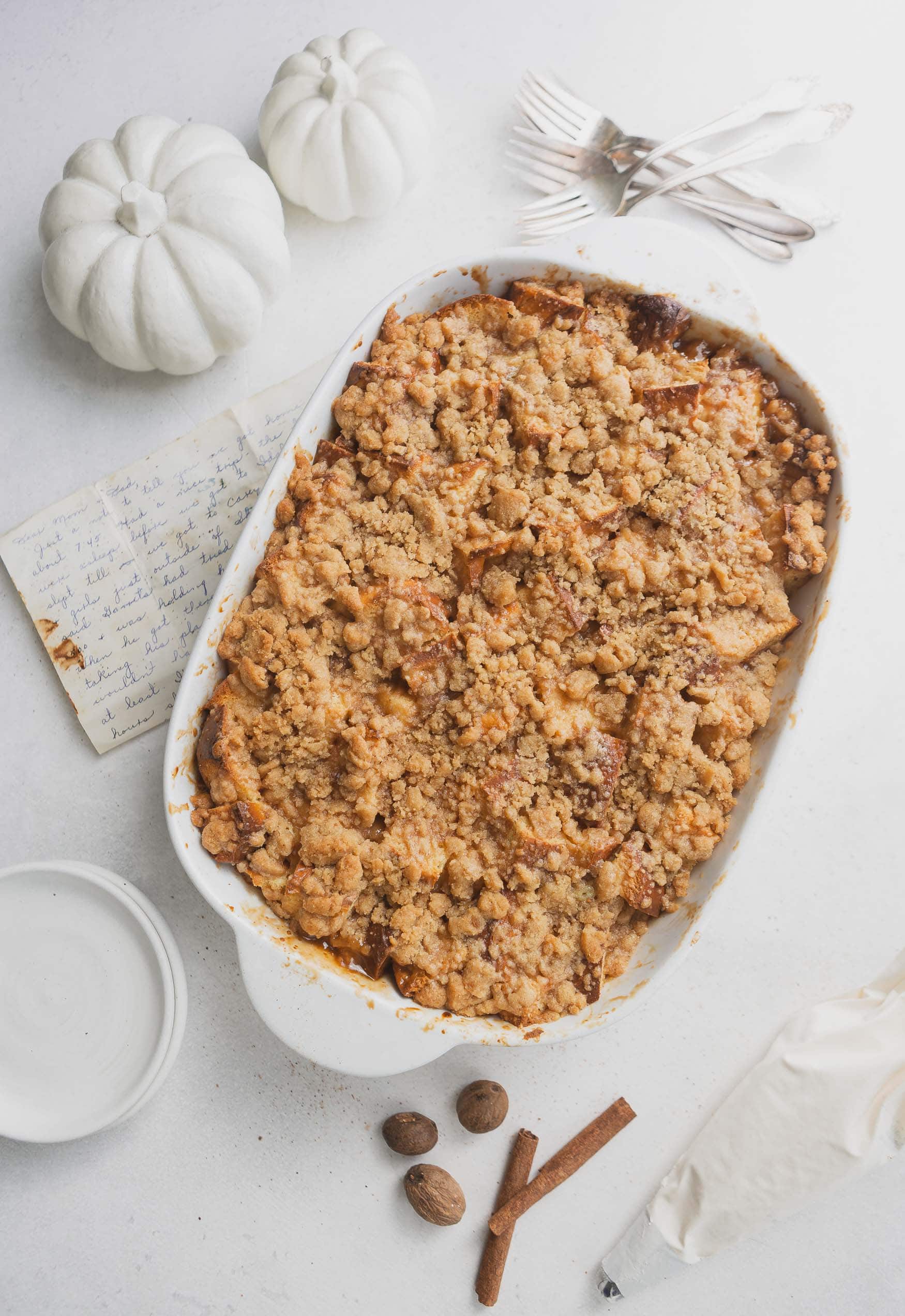A casserole dish of eggnog french toast. It is topped with a brown crumb topping. A small white pumpkin is in the background.
