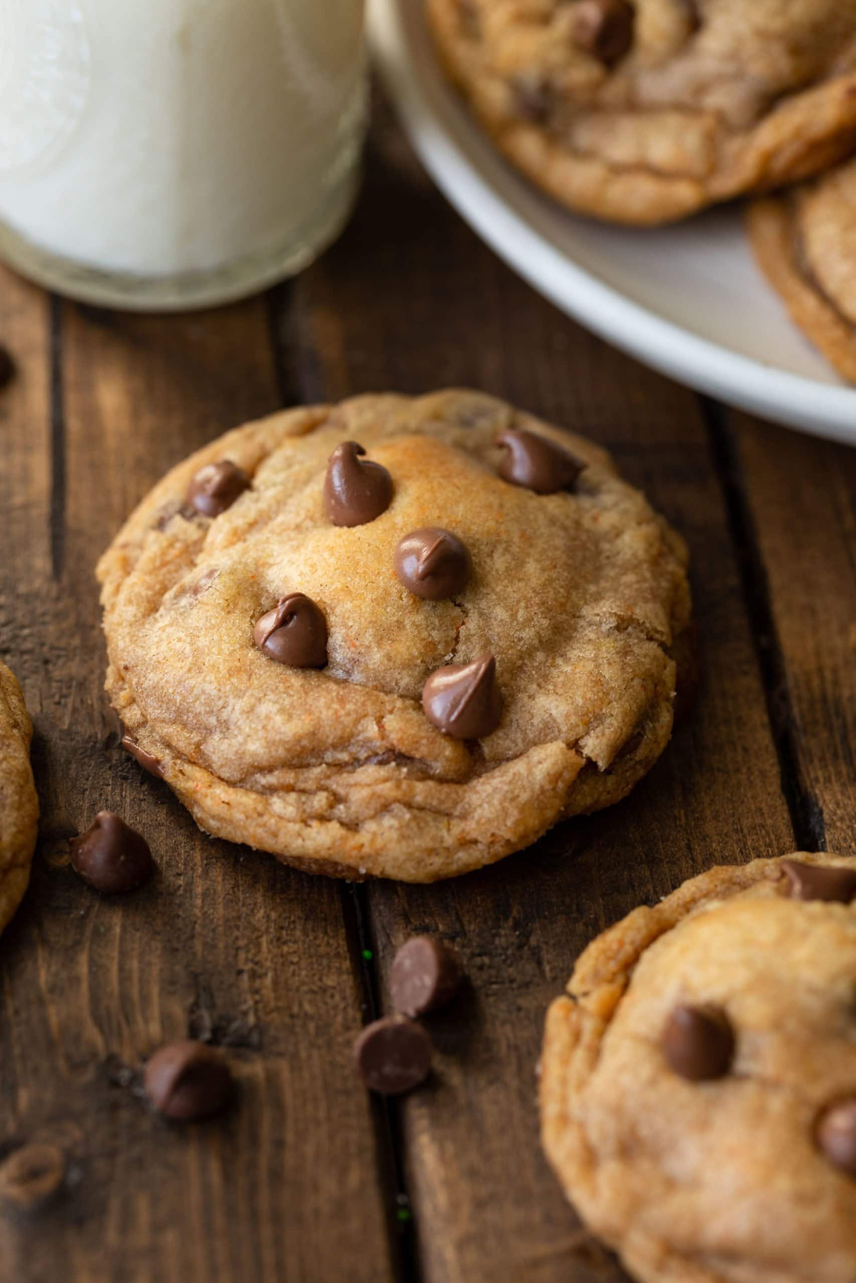 a photo of a single pumpkin pudding cookie sitting on a wooden table with chocolate chips scattered around it.