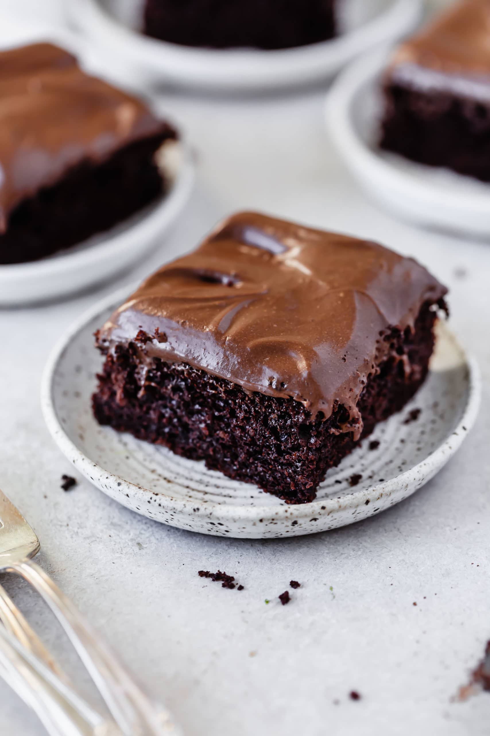 A dessert plate with a piece of chocolate mayonnaise cake frosted with chocolate cream cheese frosting. There are two more plates of cake in the background.