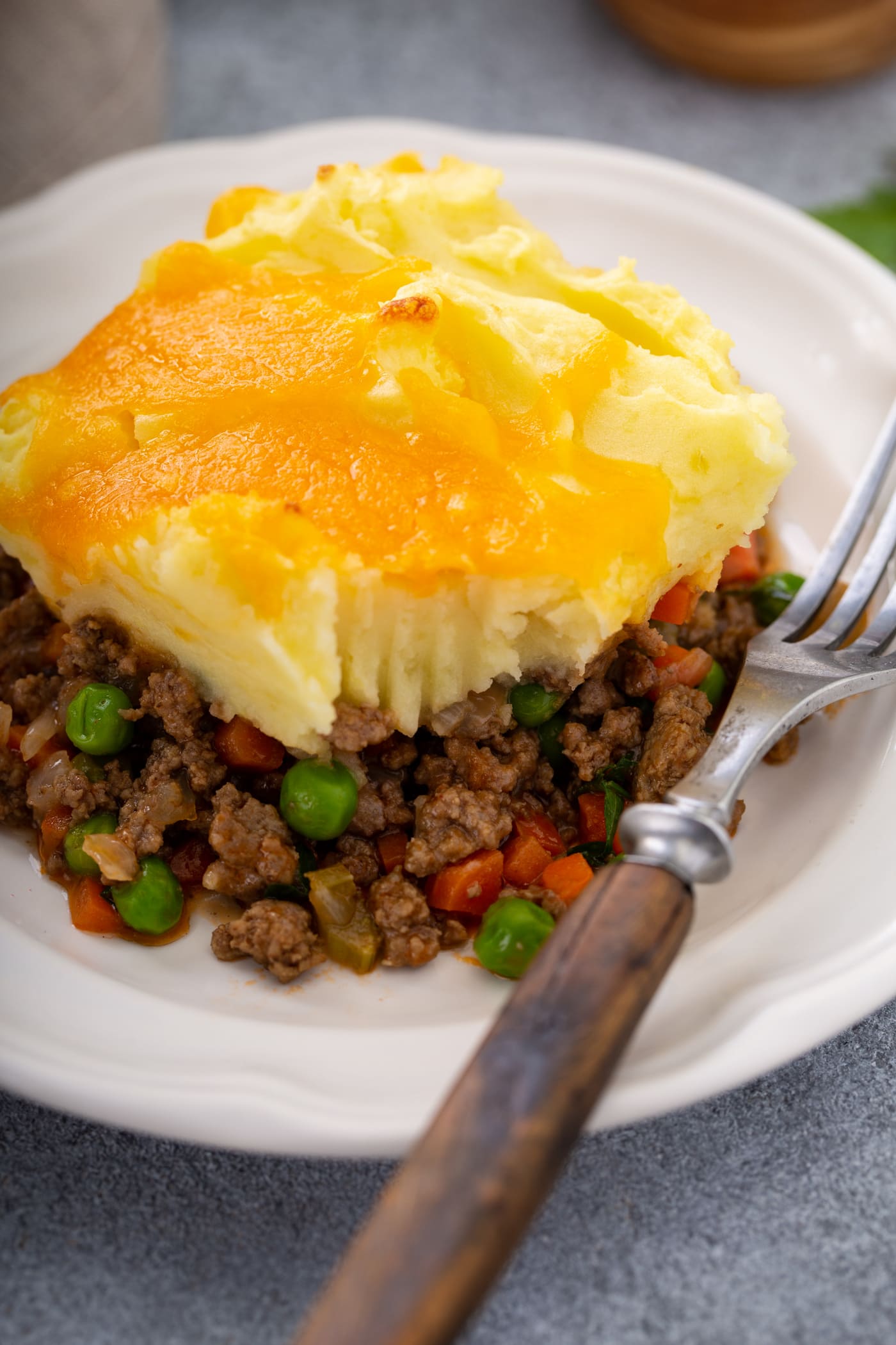 A serving of shepherd's pie on a white plate with a fork resting beside it. You can see the rich ground beef and vegetable filling, the mashed potatoes on top, and the yellow melted cheddar cheese topping it all.